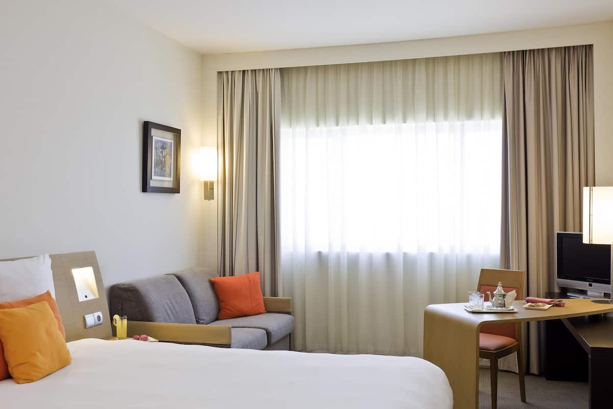 Ein Hotelzimmer mit einem großen Bett, einem Schreibtisch mit Computer, einem Sessel, einer Couch mit Kissen, einem Fenster mit Vorhängen und einer Wandleuchte.