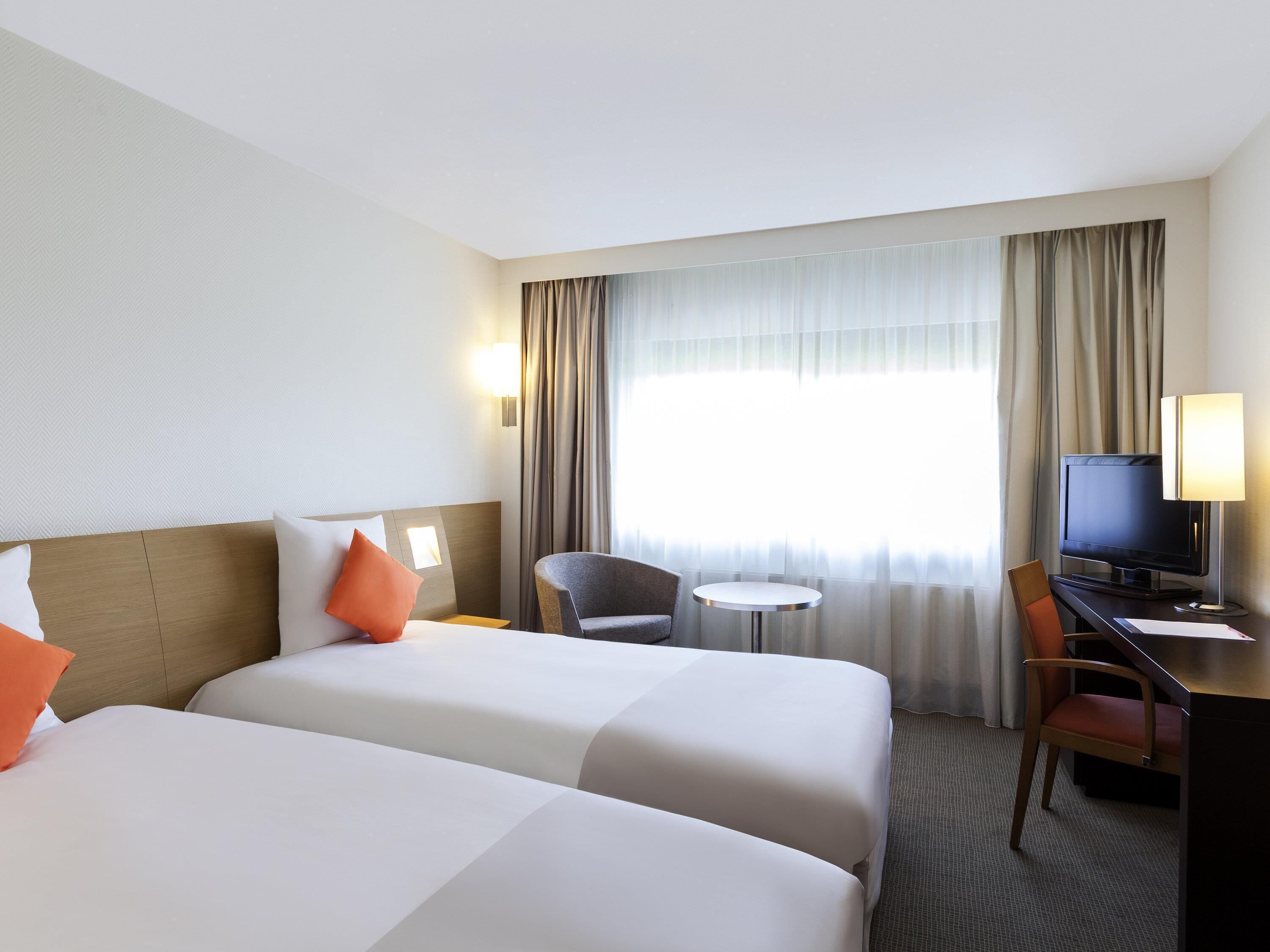 A hotel room with two beds, a desk, a chair, a television, and a window with curtains.