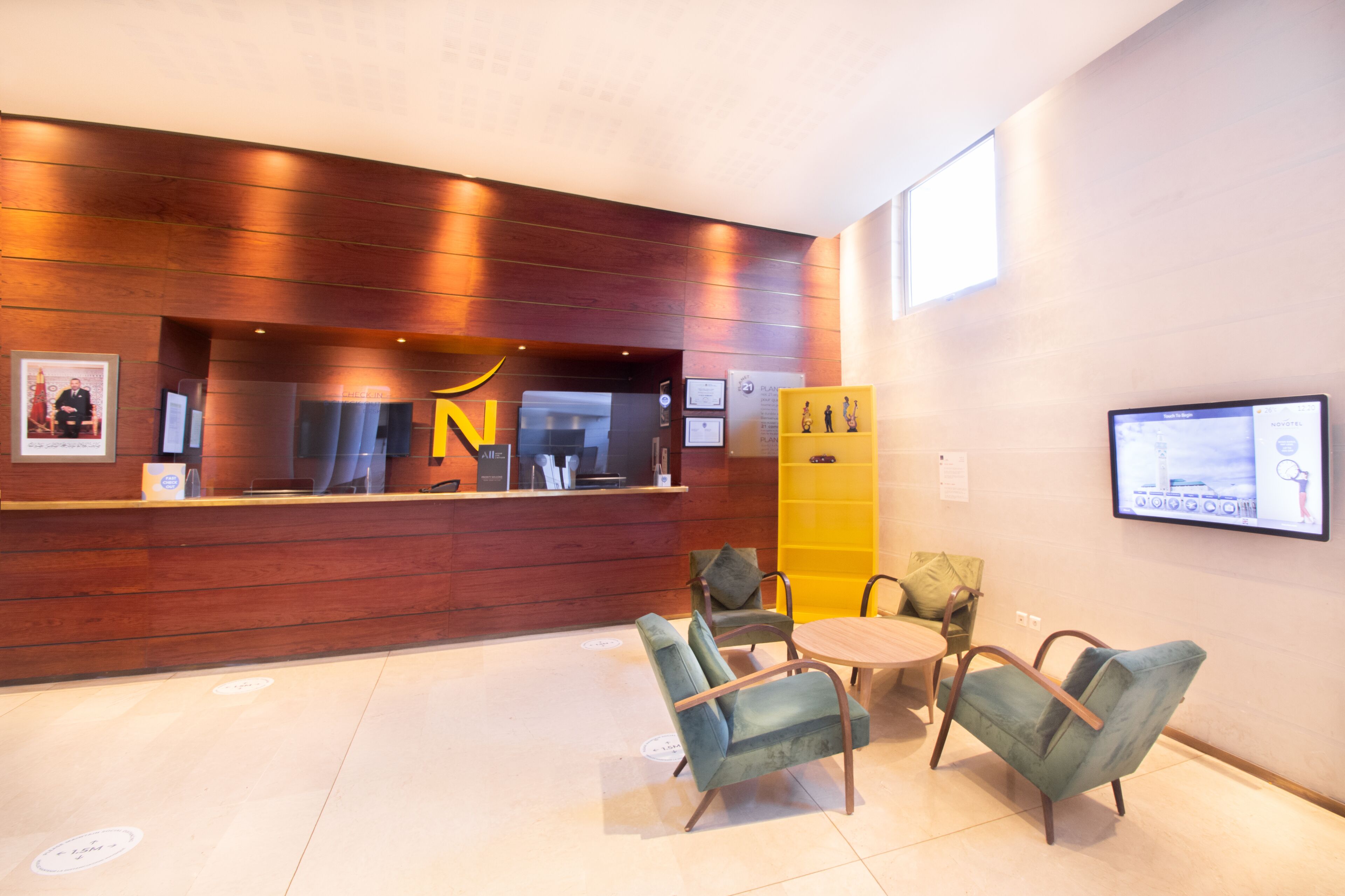Un área de recepción con un mostrador de madera, un gran logotipo 'N', un atril informativo amarillo y una pantalla de televisión en la pared.