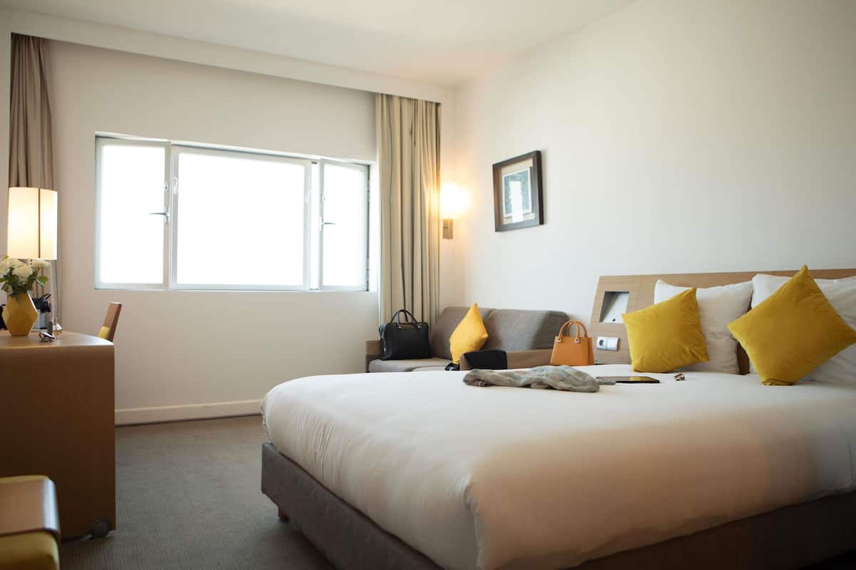 A hotel room with a large bed, a bedside table, a lamp, a window, and a wall-mounted picture.