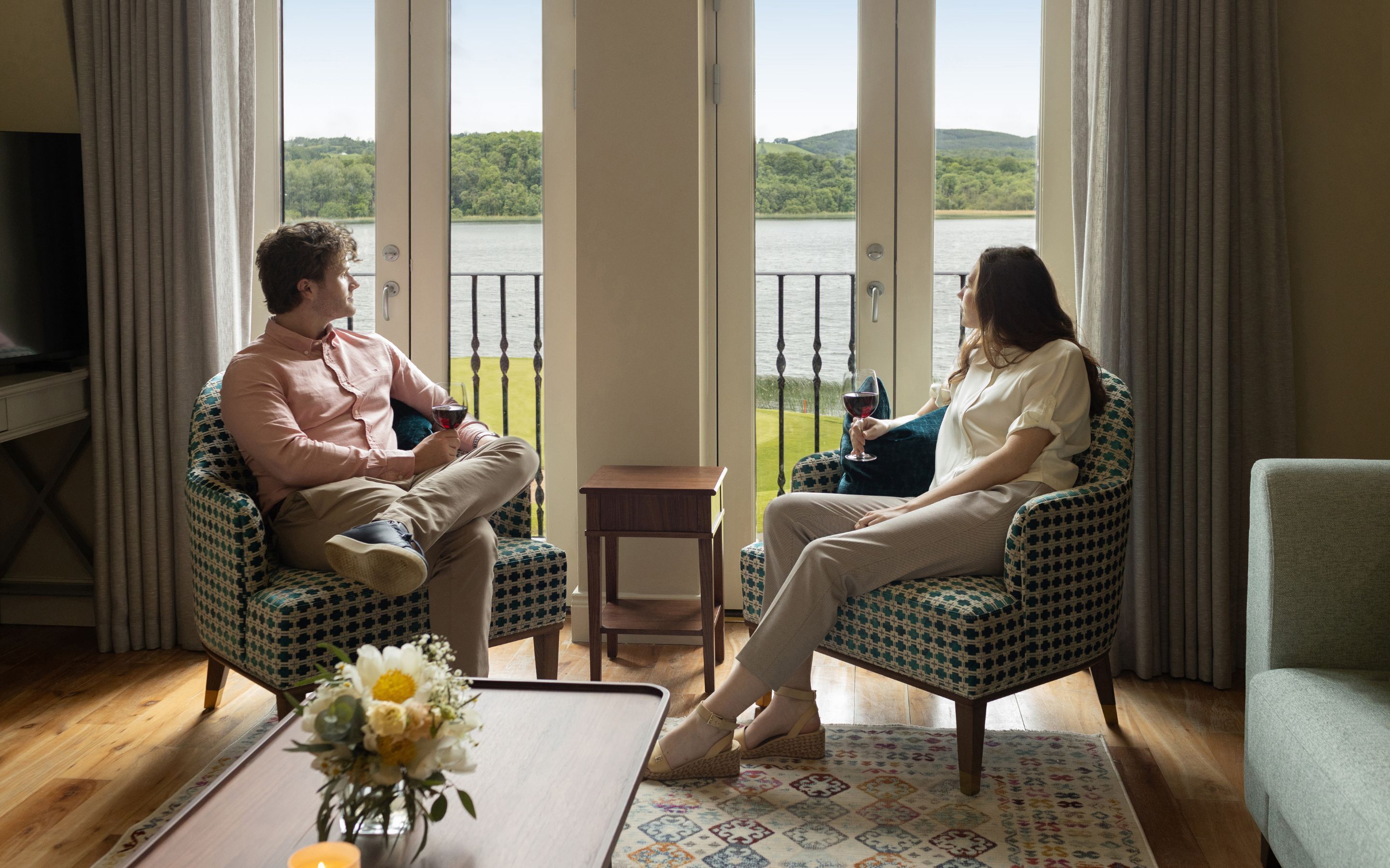 A room with a large window offering a view of a lake and hills. There are two chairs facing each other, a small table with a vase of flowers, and a television on a stand.