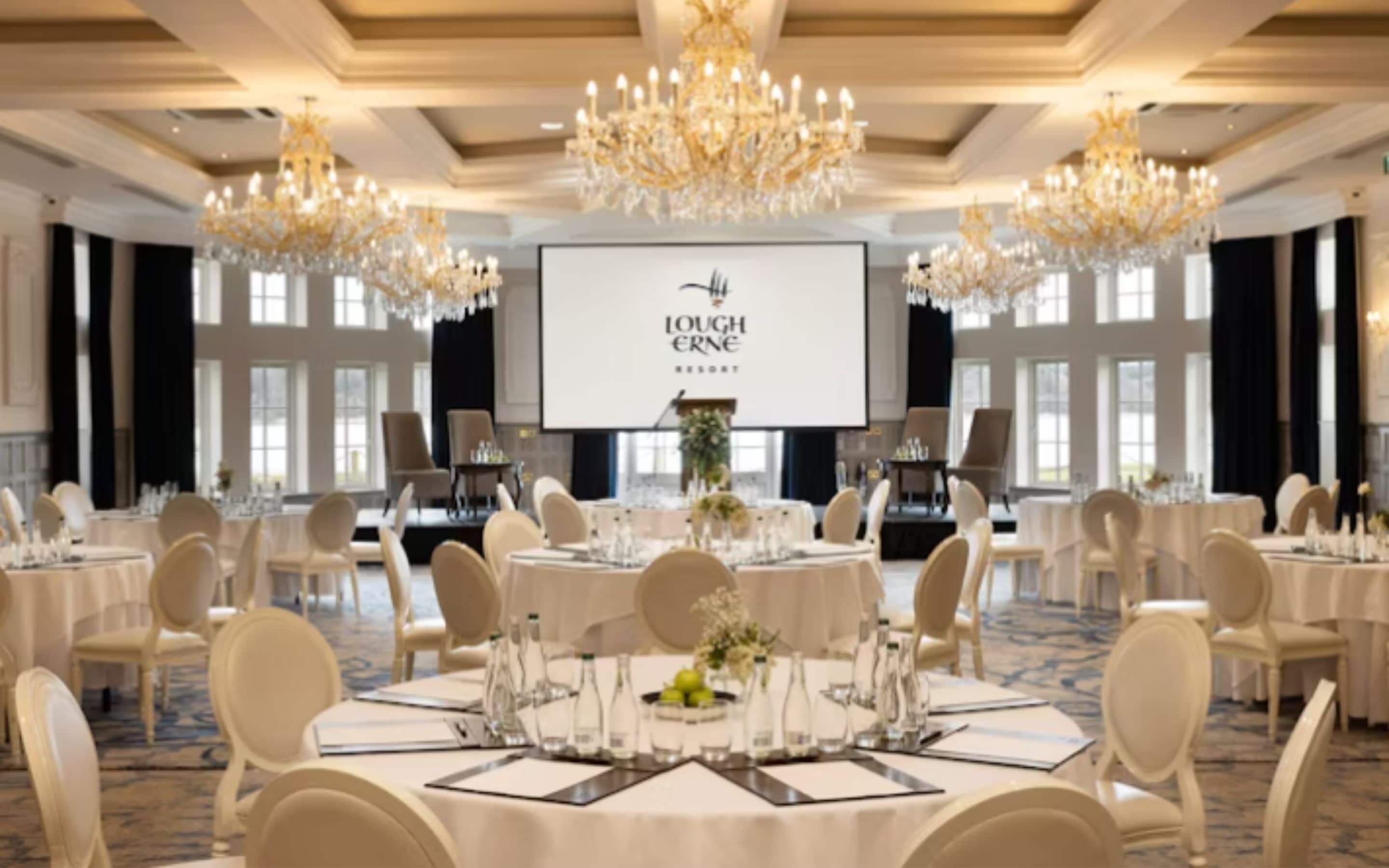 En elegant festlokal på Lough Erne Resort med runda bord uppdukade för middag, kristallkronor och en projektionsduk som visar resortens namn.