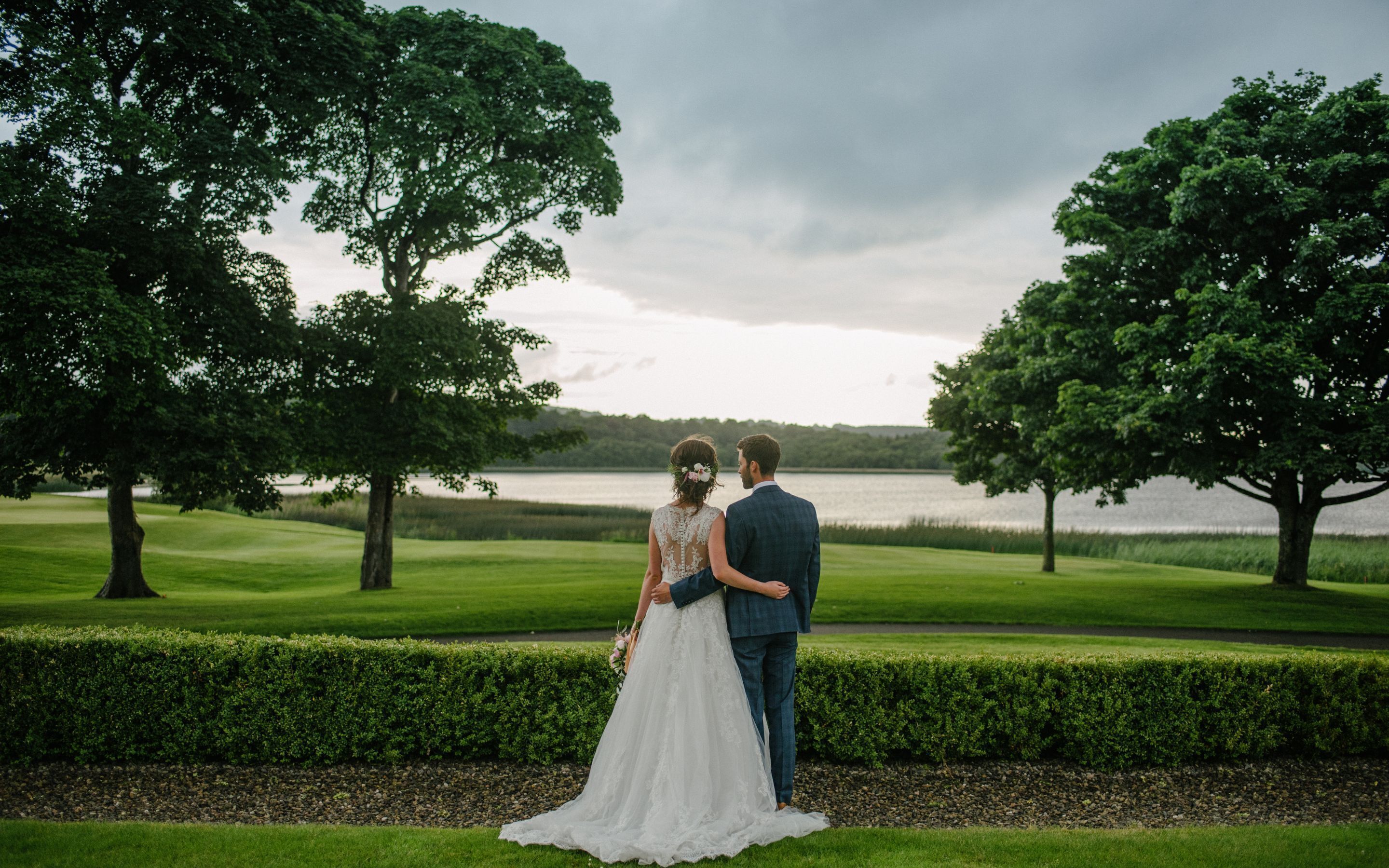 A couple standing in a lush, green landscape with a lake in the background.