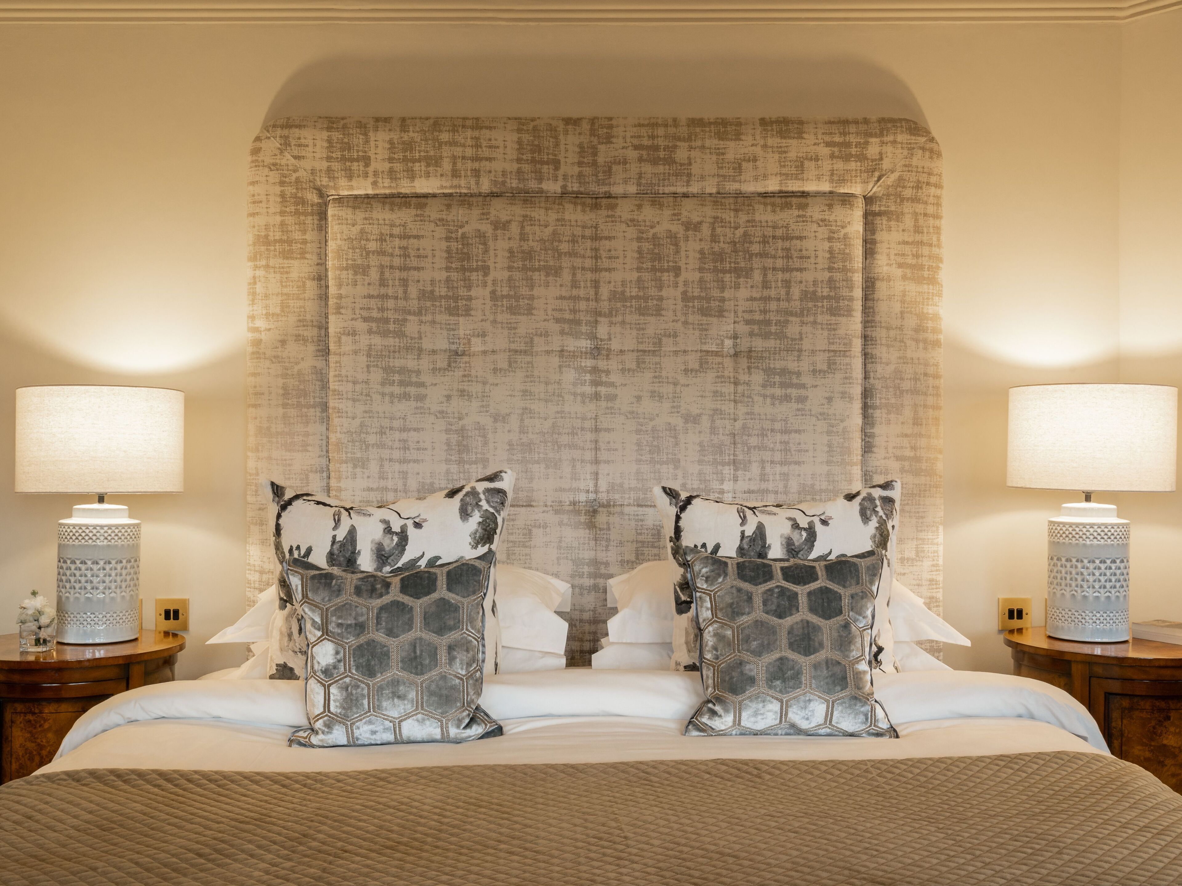 Habitación de hotel con una cama grande, dos almohadones decorativos a juego, dos lámparas de noche y un cabecero texturizado.