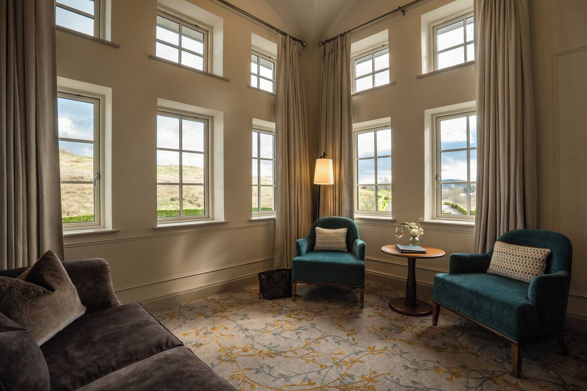 A room with a large window, a green armchair, a brown sofa, and a small table with a vase.