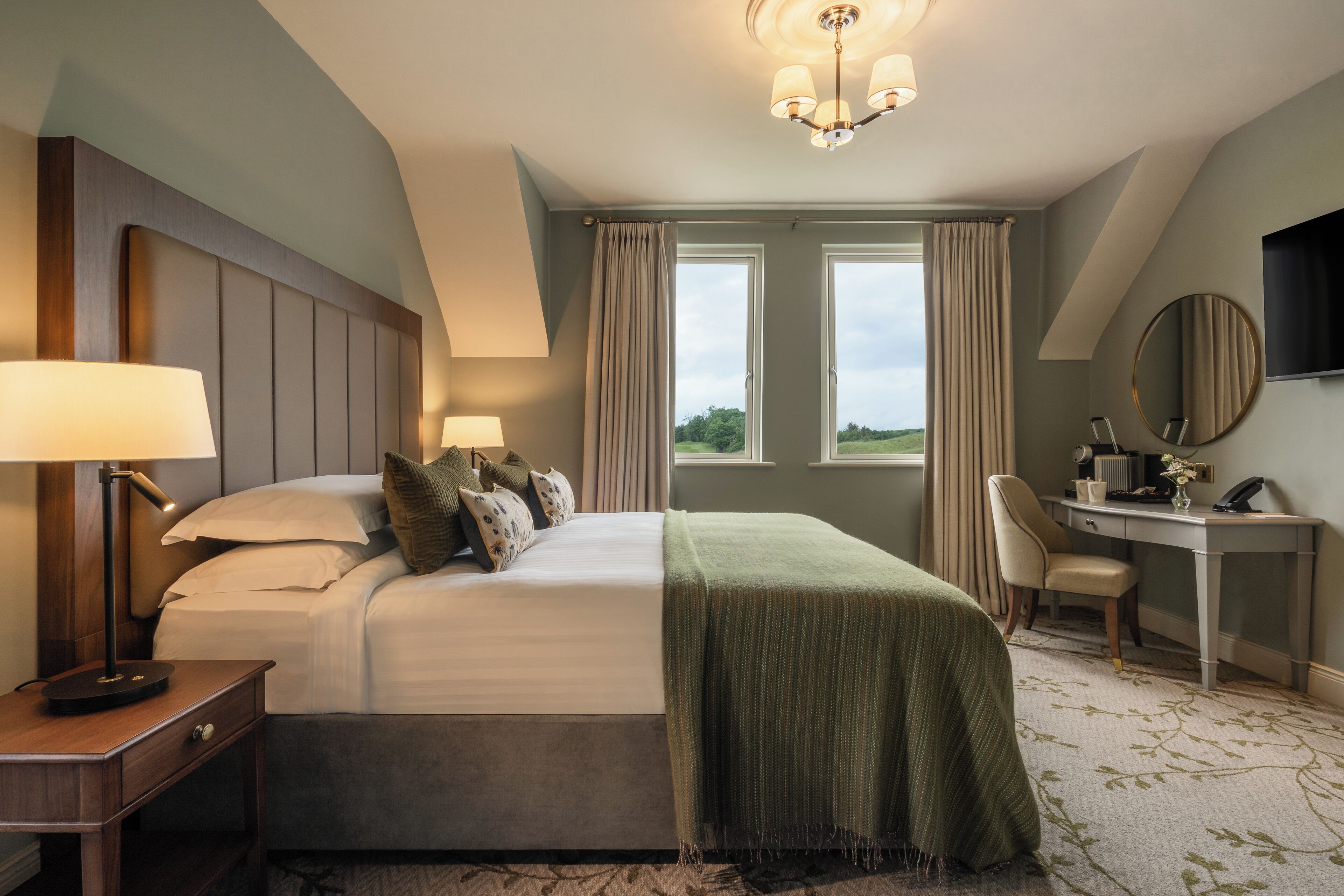 A hotel room with a large bed, two bedside lamps, a desk with a chair, a round mirror, and a view of the outdoors through two windows.