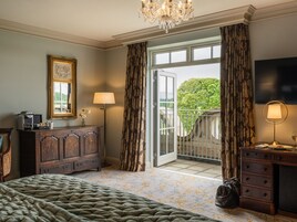 Habitación de hotel con cama, cómoda, escritorio, lámpara, televisión y balcón con vista a árboles.