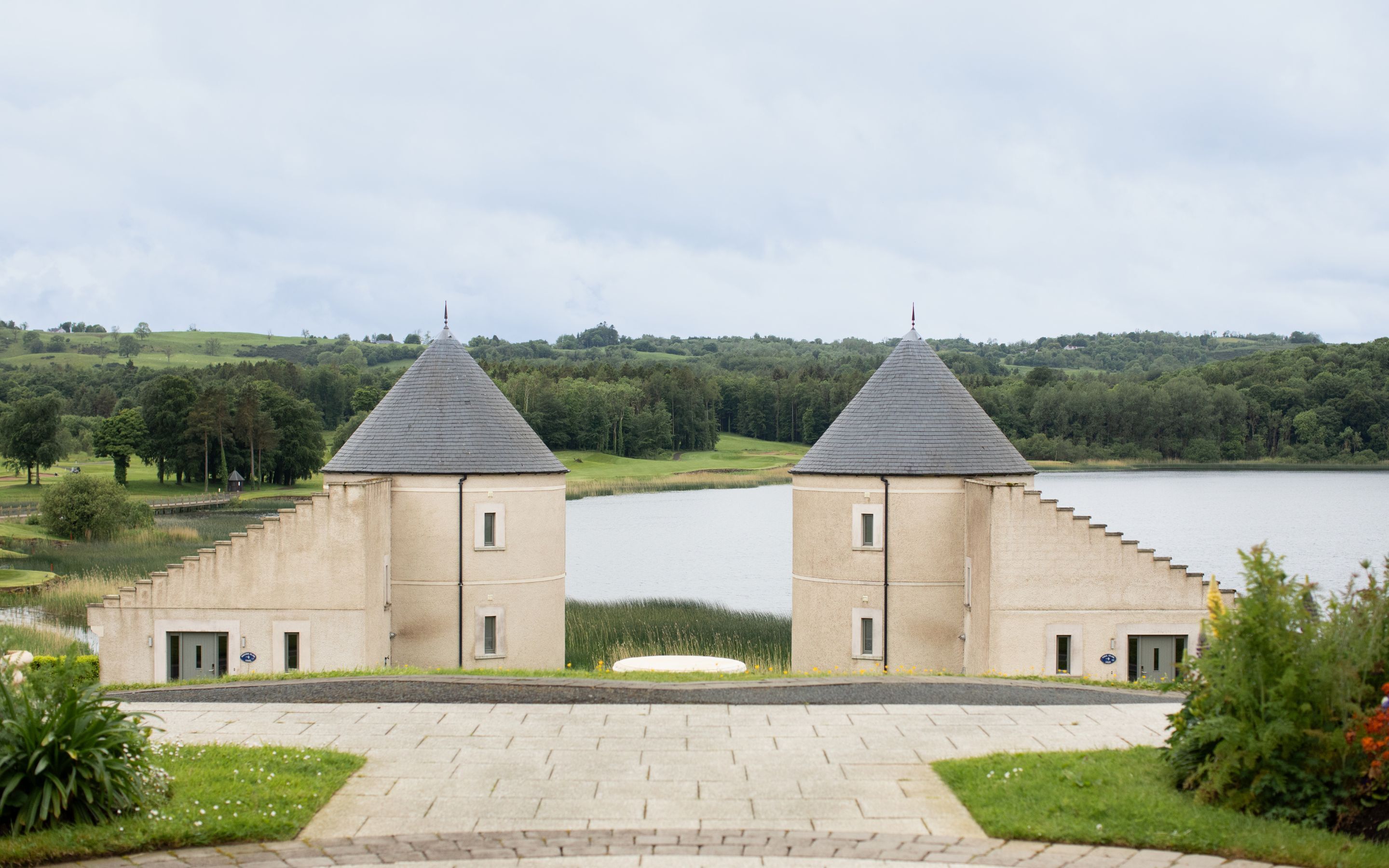 A symmetrical building with two towers, a paved entrance, and a lake in the background.