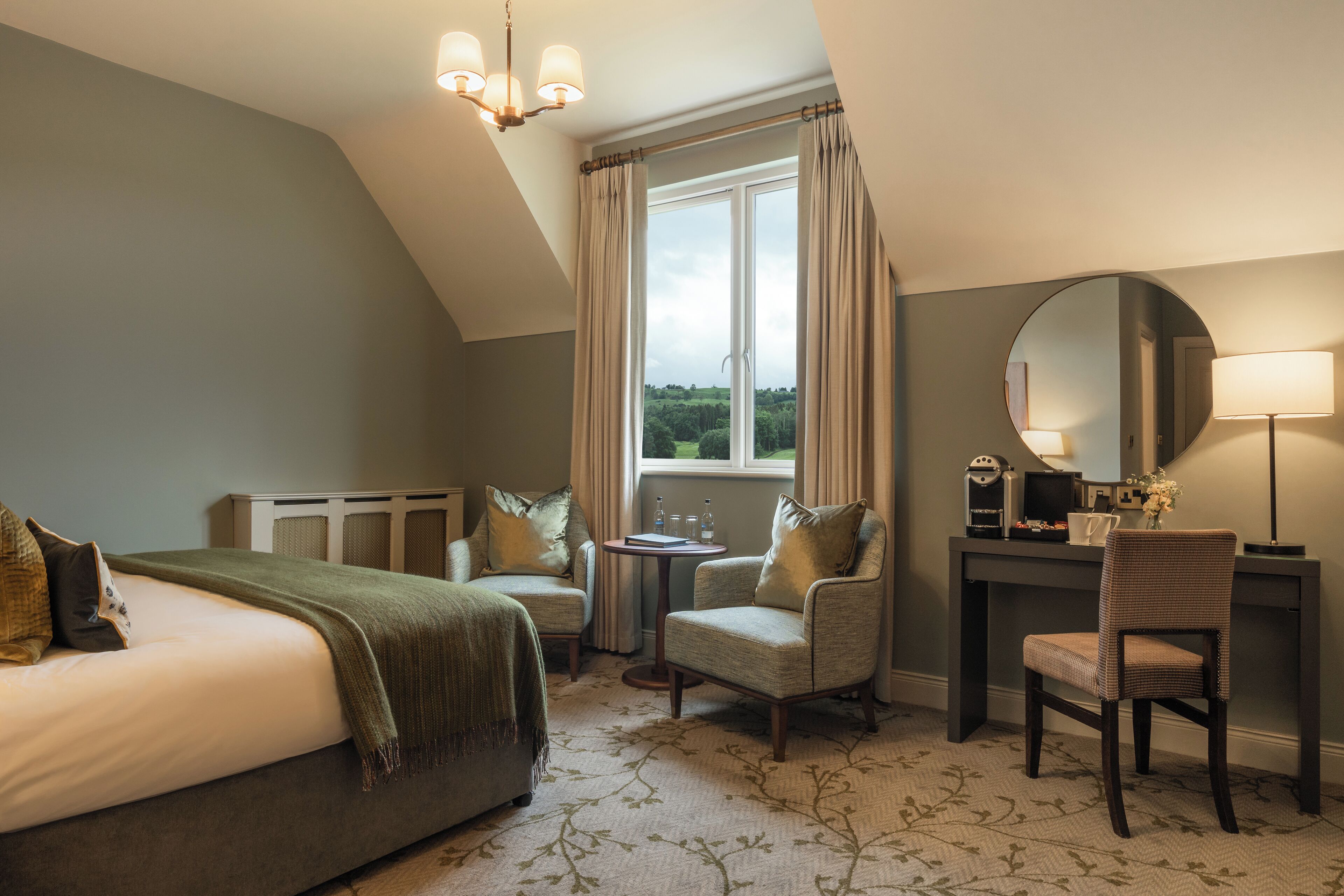 A bedroom with a bed, two armchairs, a desk, a mirror, and a window with curtains.