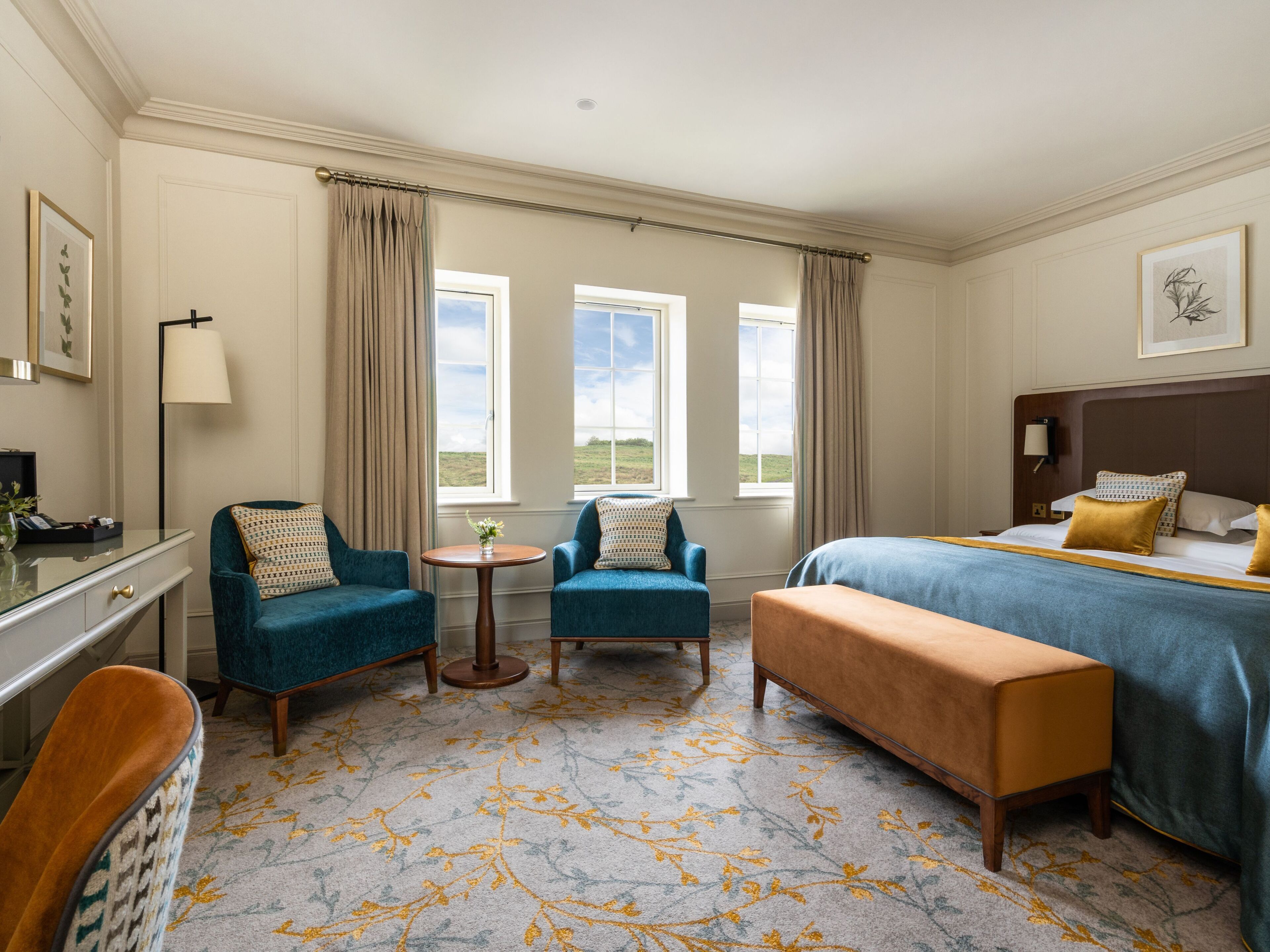 A hotel room with a bed, two armchairs, a small table, a desk, and a chair. There are windows with curtains, a lamp, and artwork on the walls.