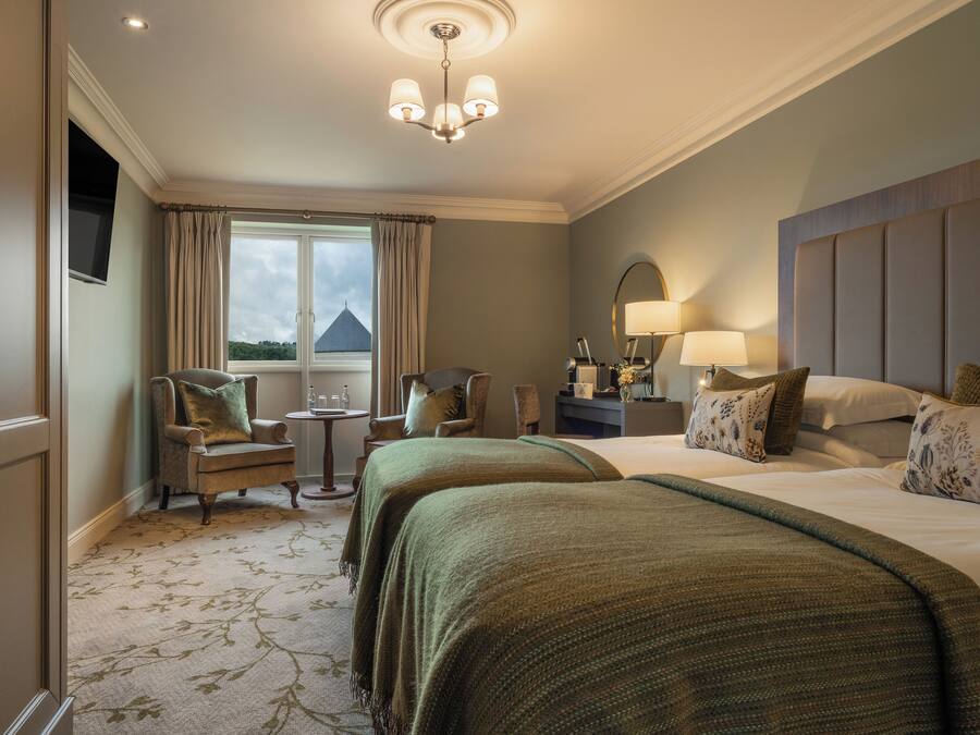 Habitación de hotel con dos camas, zona de estar con sillas, una mesita, ventana con cortinas y un candelabro.
