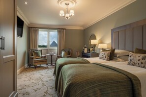 A hotel room with two beds, a sitting area with chairs, a small table, a window with curtains, and a chandelier. - Lough Erne Resort (Enniskillen)