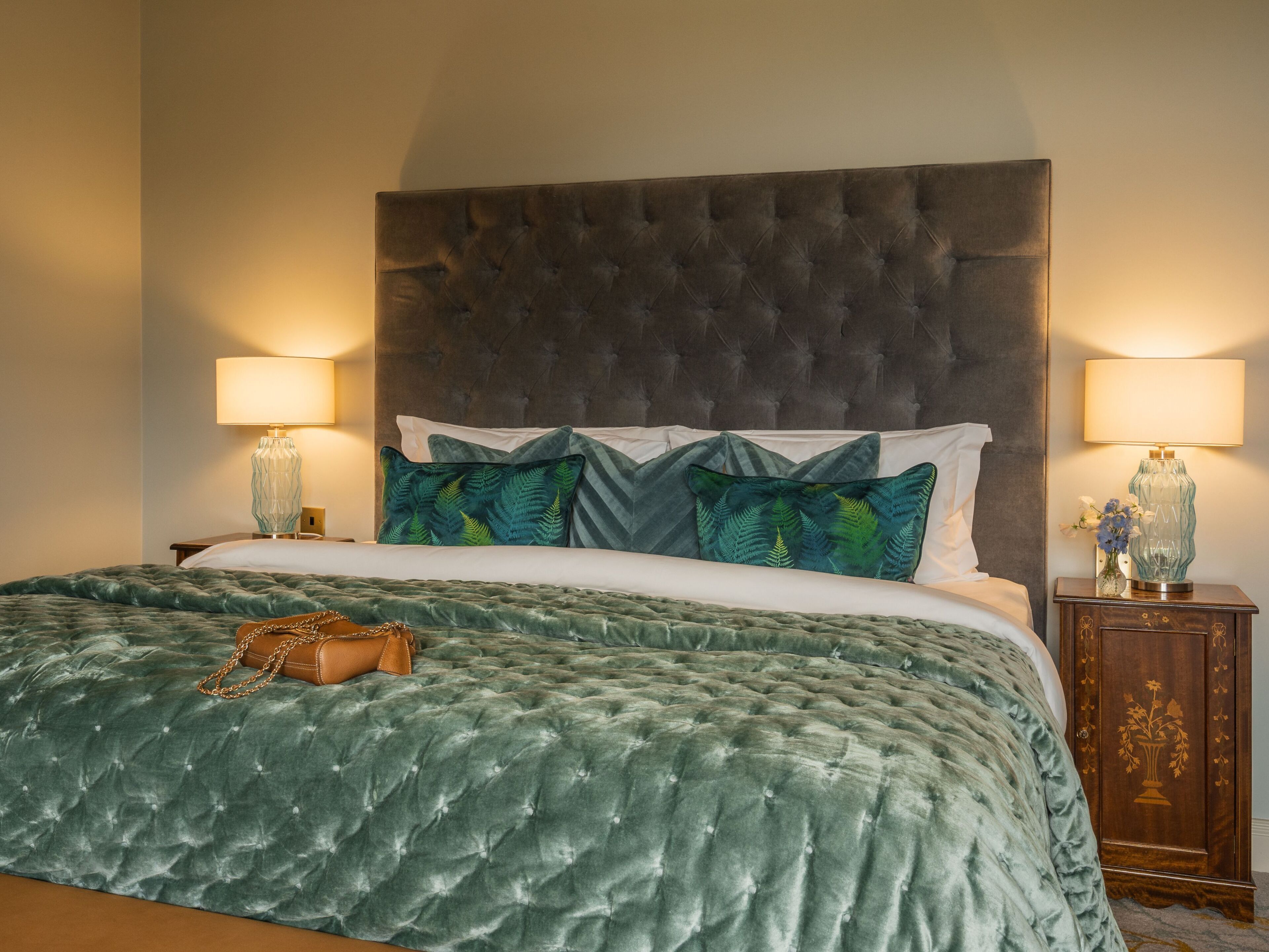 Cama bem arrumada com um espalhadeira de veludo verde, duas almofadas decorativas e uma bolsa marrom sobre a cama. Há duas mesas de cabeceira com luminárias e um pequeno armário de madeira com um desenho floral.