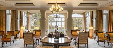 A spacious room with a large round table surrounded by chairs, large windows with curtains, and a chandelier.