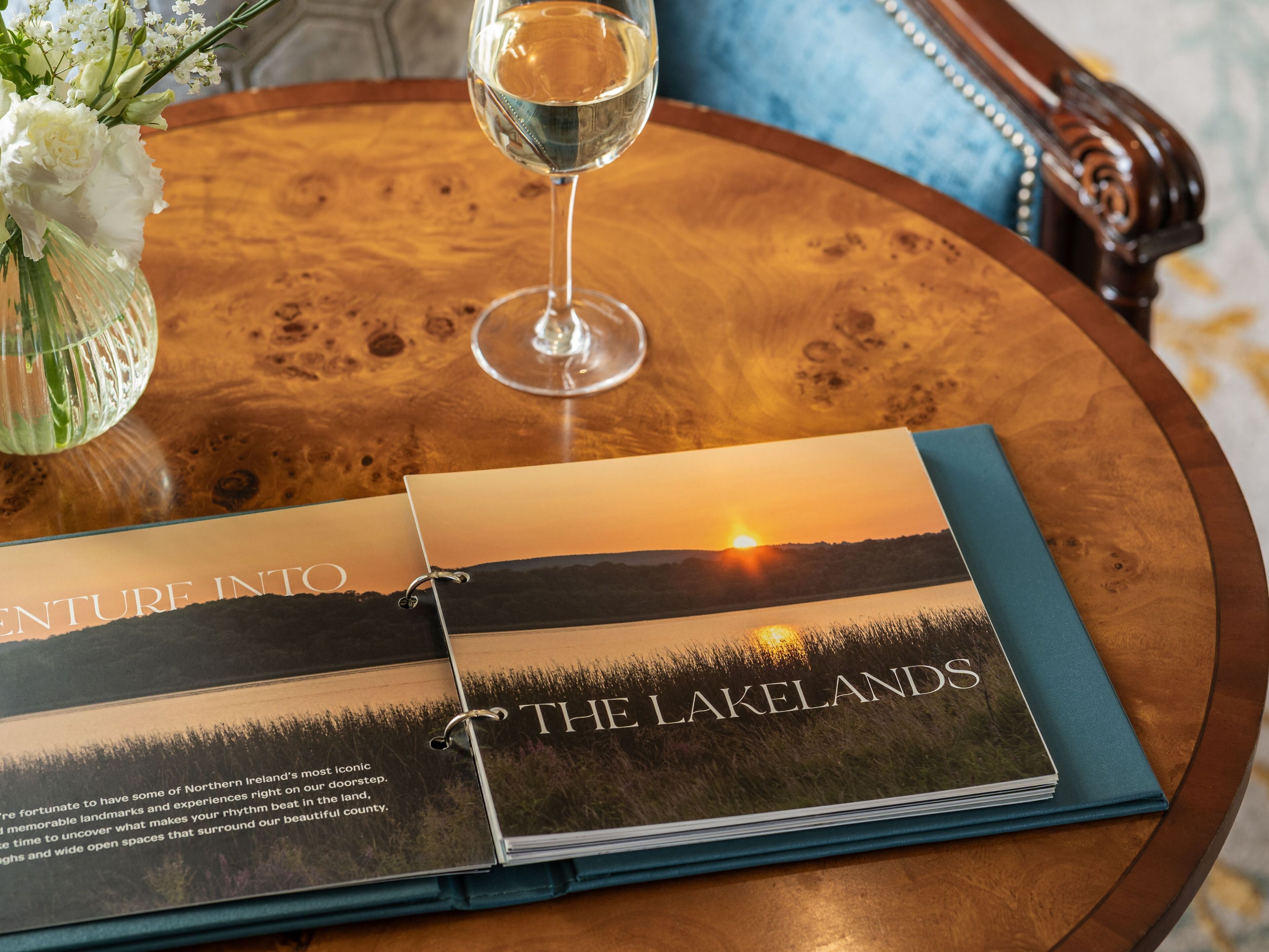 Ein Buch mit dem Titel „The Lakelands“ auf einem Holztisch zusammen mit einem Glas Wasser und einer Blumenvase.