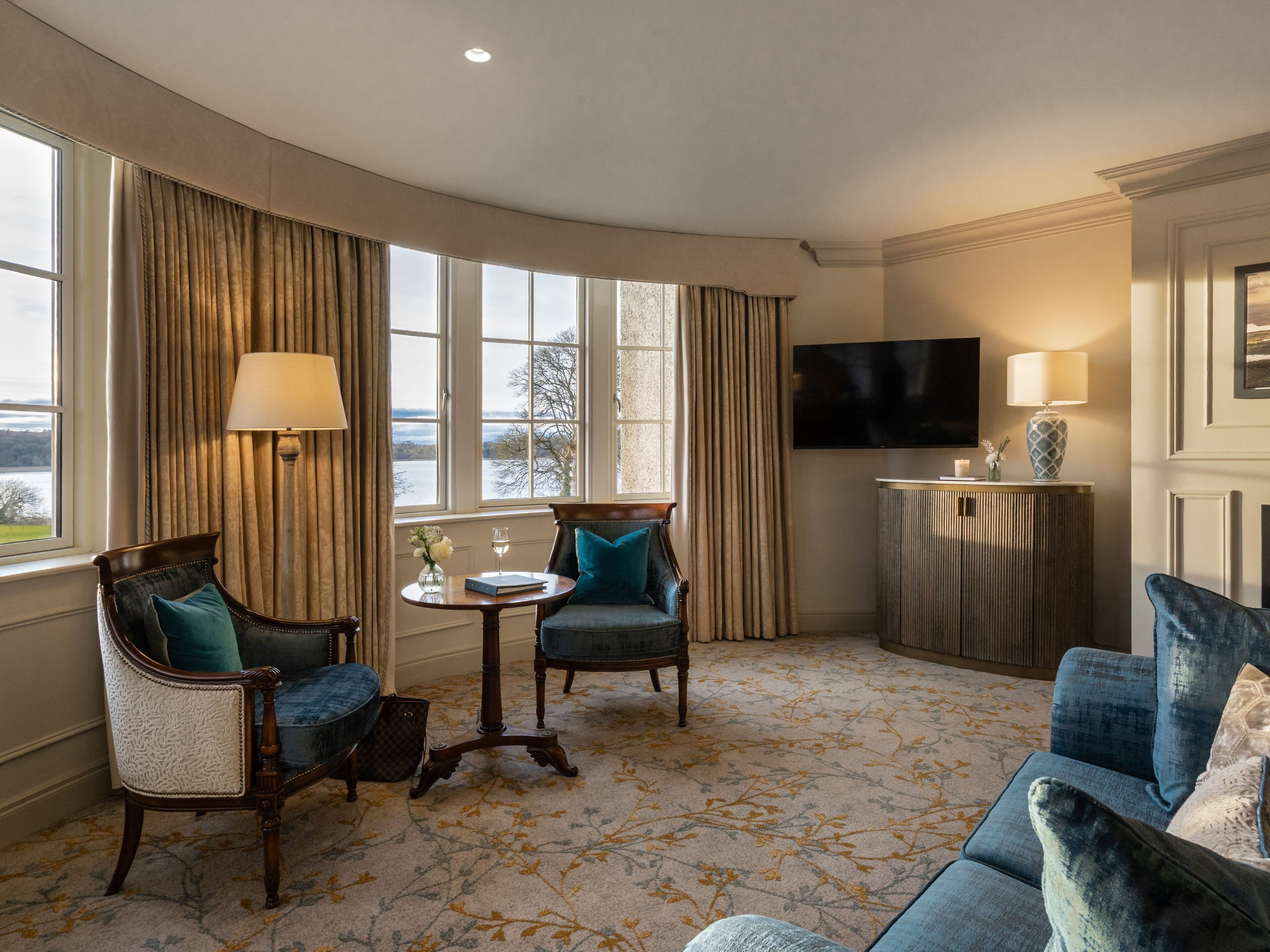 A hotel room with a curved layout, featuring a sofa, two armchairs, a small round table, a TV, and a view of the outdoors through large windows.