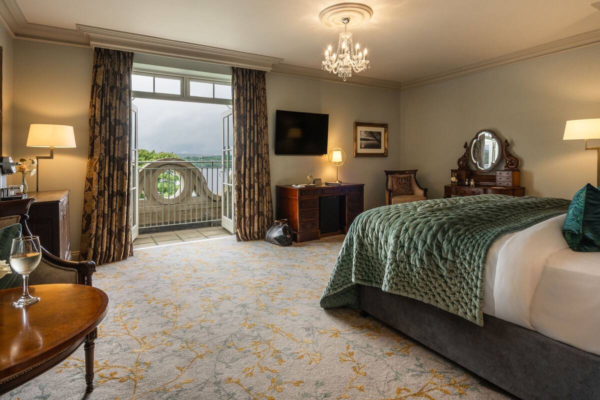 A hotel room with a large bed, a desk with a lamp, a chair, a dresser, a mirror, and a balcony with curtains.