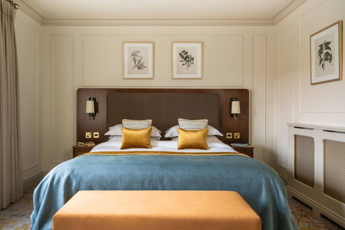A hotel room with a large bed, two bedside tables, a nightstand, a bench, and framed botanical prints on the wall.