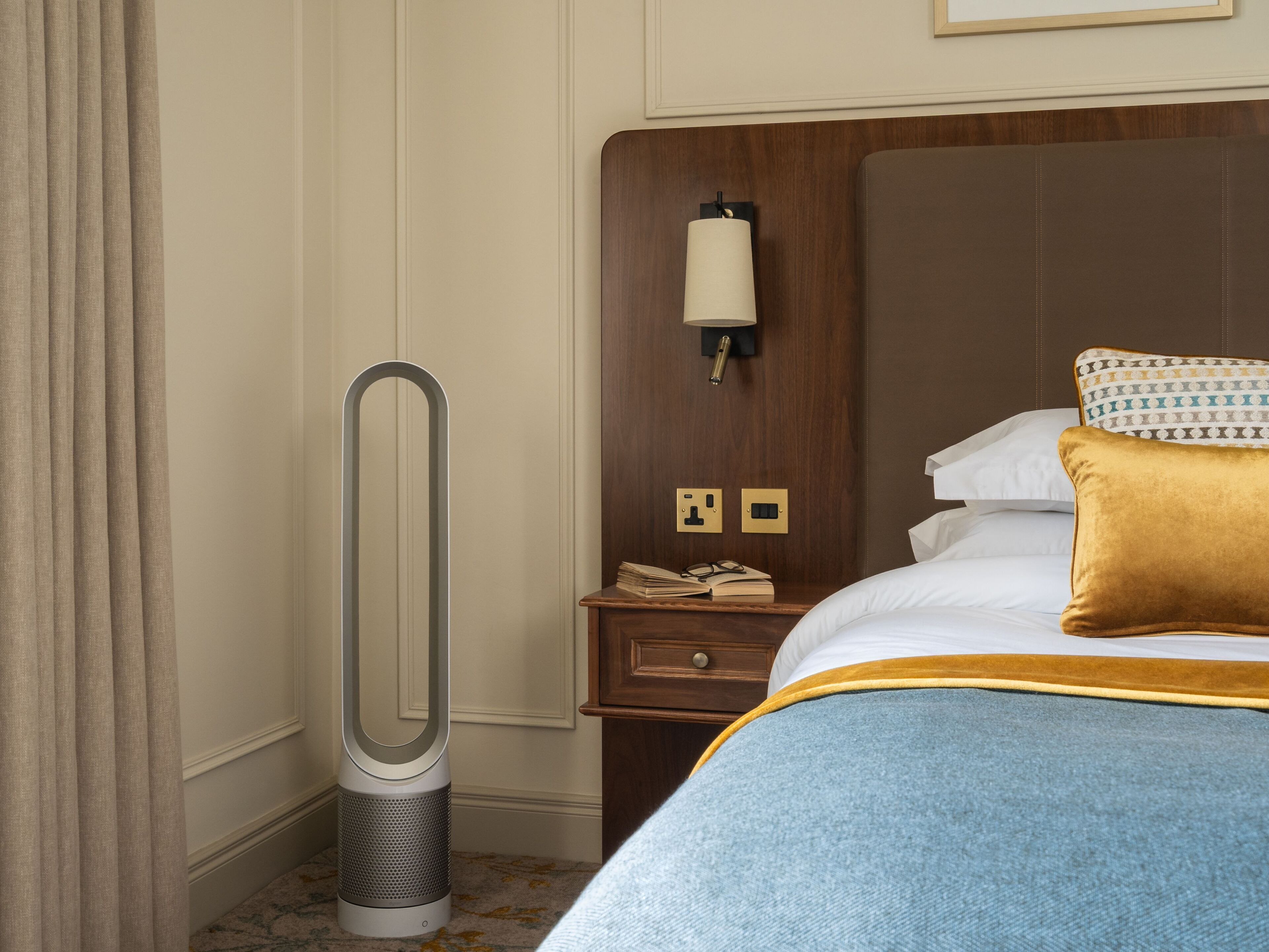 A hotel room with a bed, a bedside table, a nightstand, a lamp, and a standing heater.