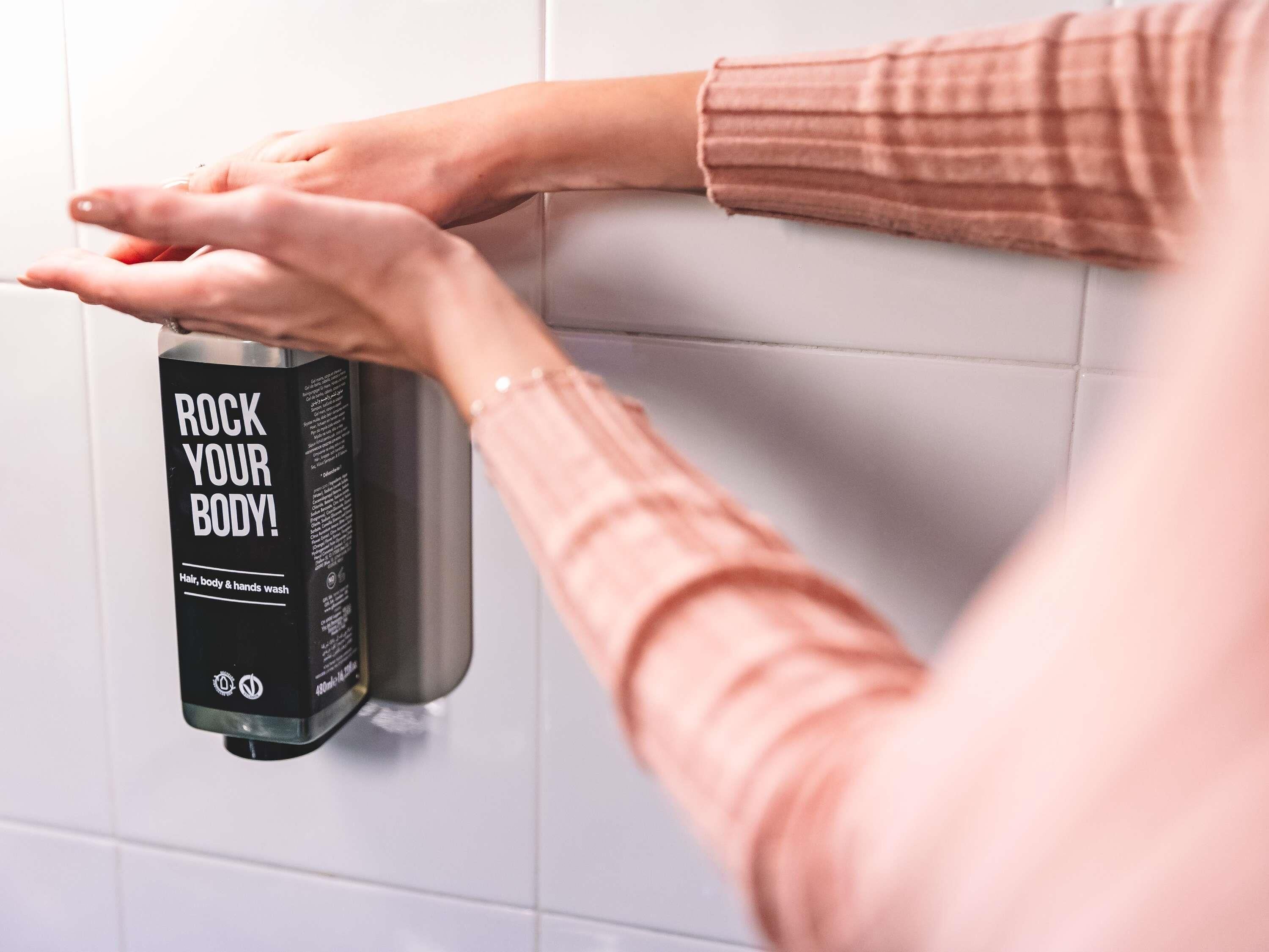 Uma pessoa utilizando um dispensador de álcool em gel fixado na parede, com etiqueta que diz 'ROCK YOUR BODY!'.