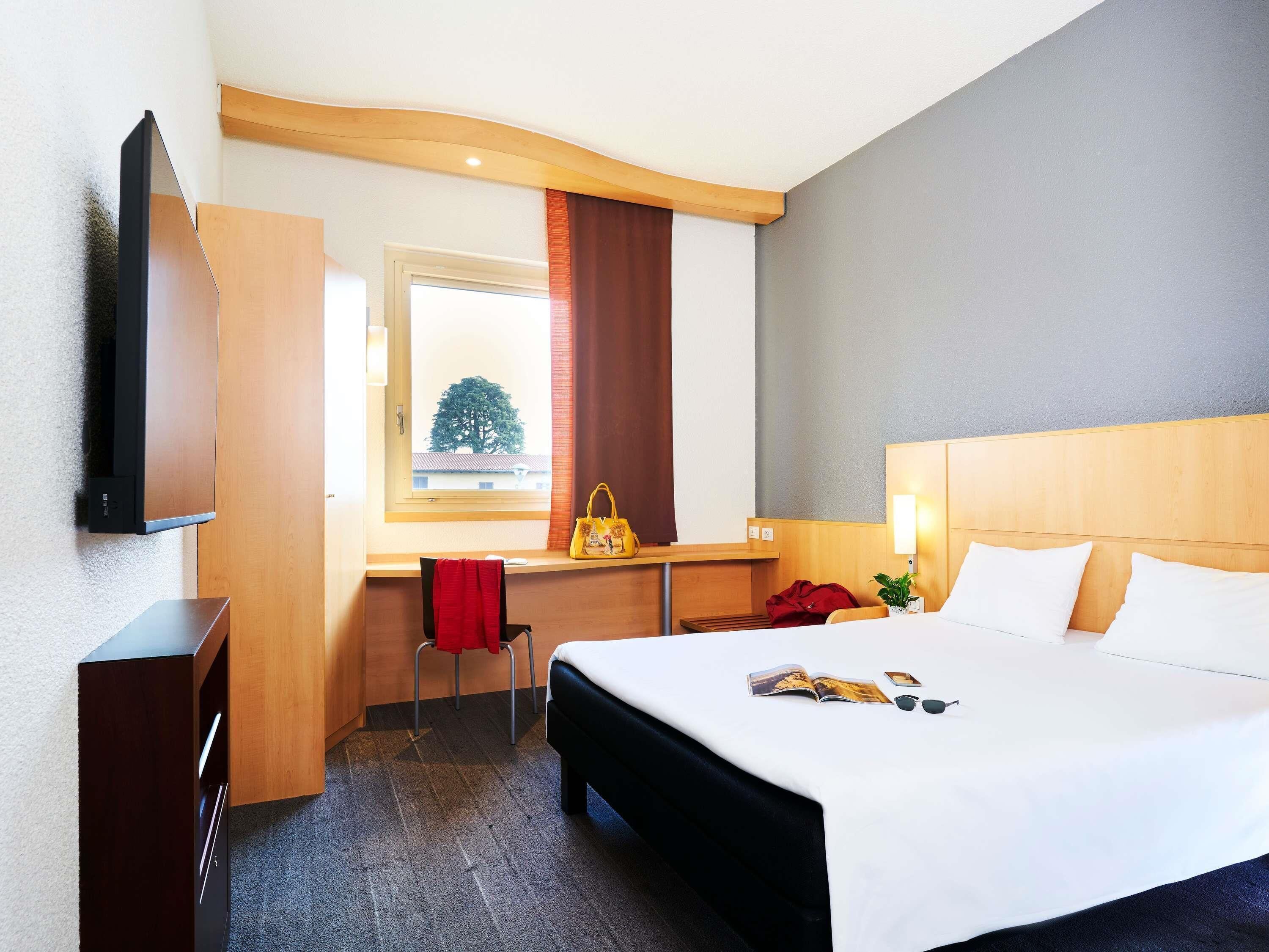 Ein Hotelzimmer mit Bett, Nachttisch, Fernseher, Schreibtisch, Stuhl und Blick auf einen Baum.
