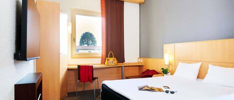 Ein Hotelzimmer mit Bett, Nachttisch, Fernseher, Schreibtisch, Stuhl und Blick auf einen Baum.