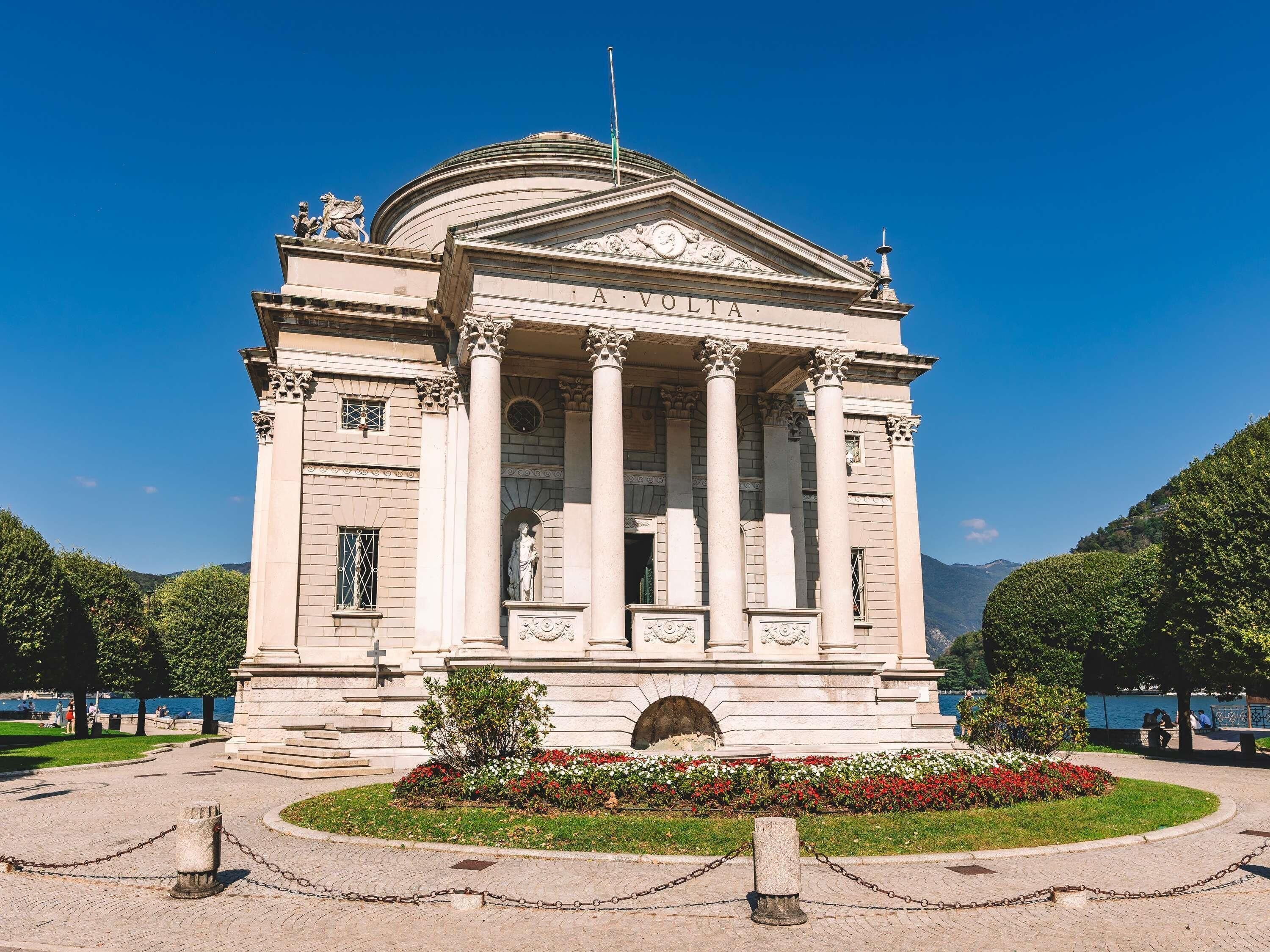 Un edificio neoclásico con columnas y una cúpula, rodeado de un jardín ajardinado y una extensión de agua.