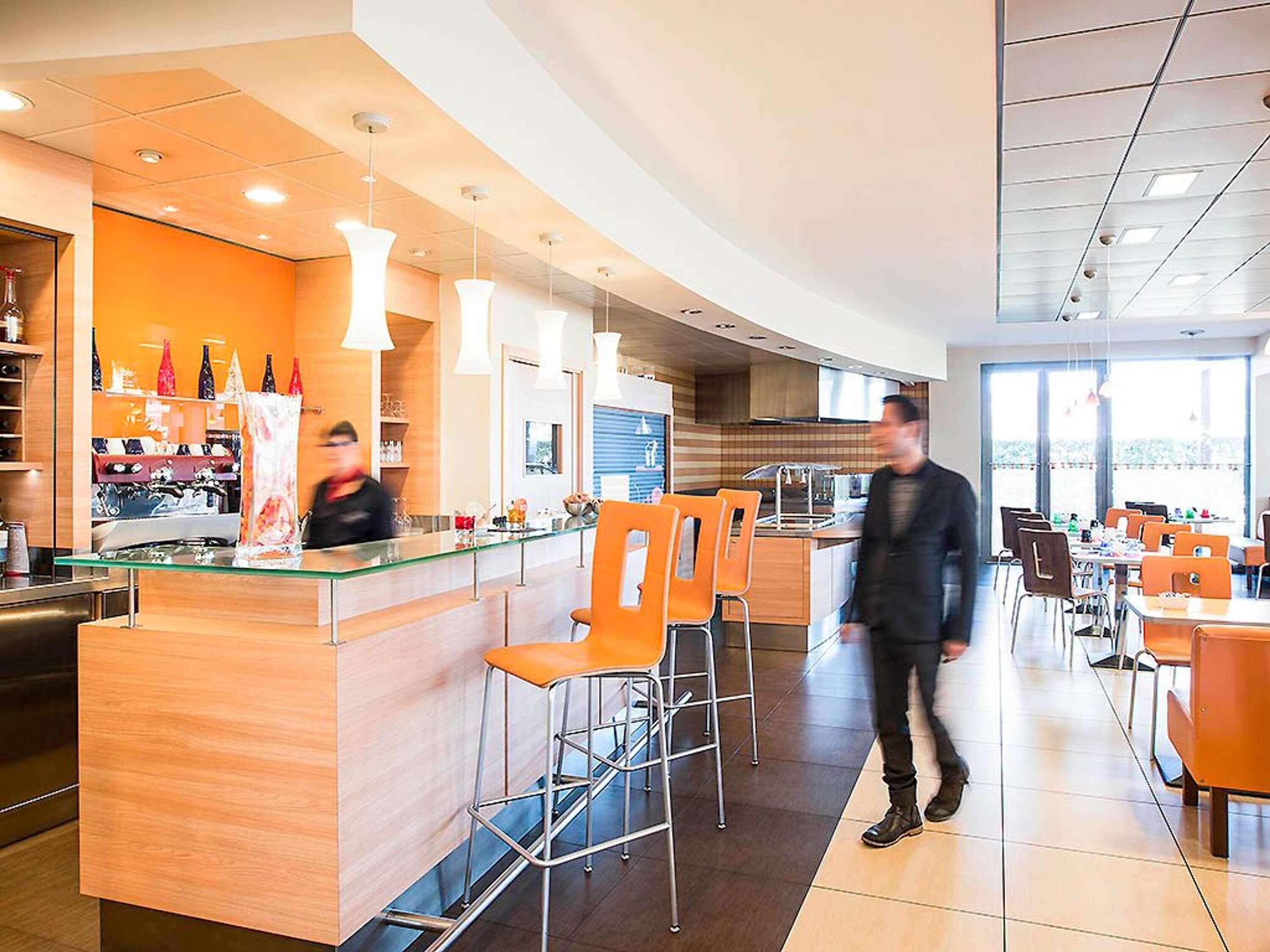 A modern restaurant with a central bar, orange chairs, and a person walking.