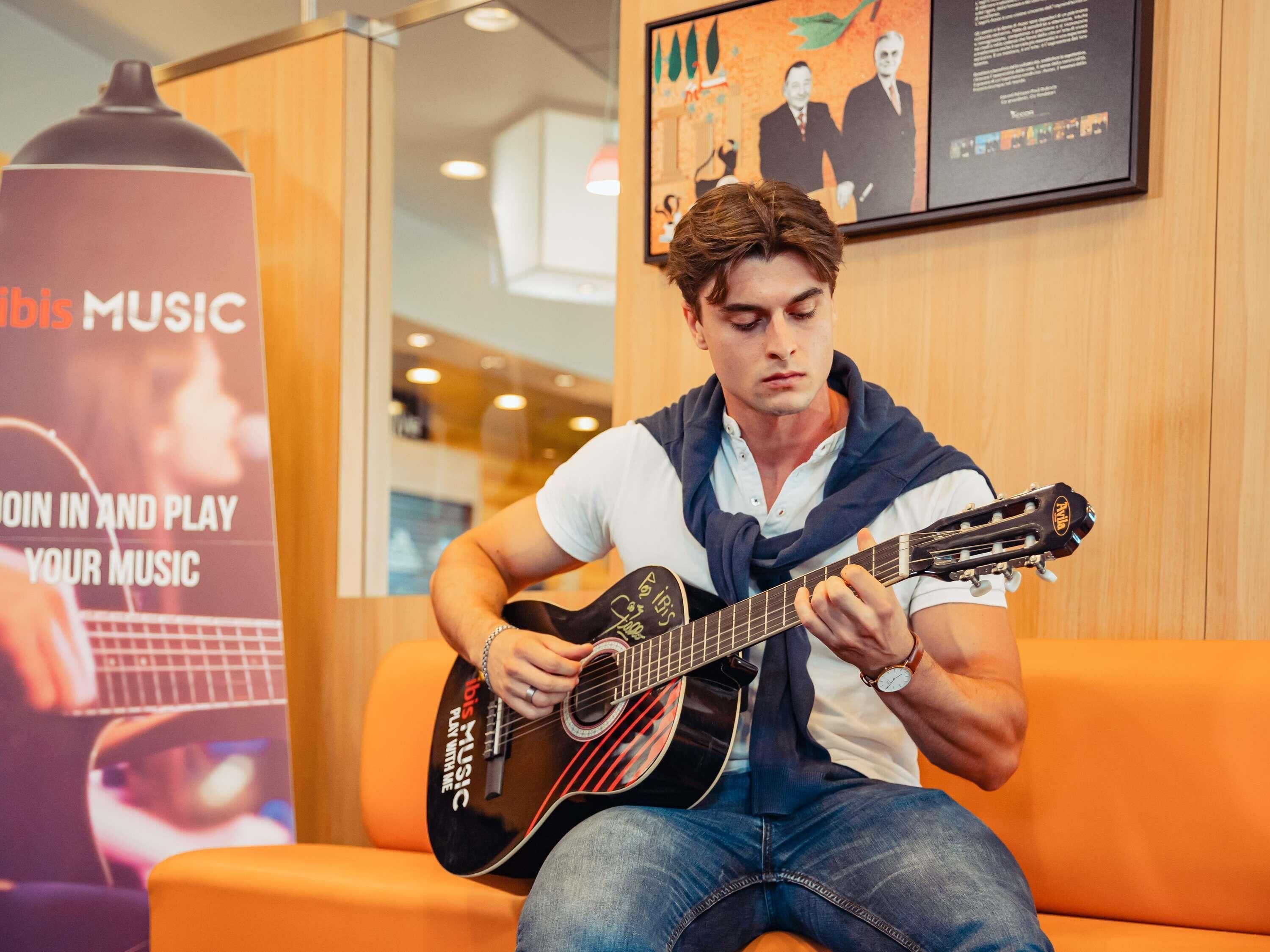Eine Person spielt Gitarre in einem Raum mit einem orangefarbenen Sofa und einem Poster, das 'Ibiza Music' anwirbt.
