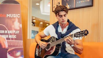 Eine Person spielt Gitarre in einem Raum mit einem orangefarbenen Sofa und einem Poster, das 'Ibiza Music' anwirbt.
