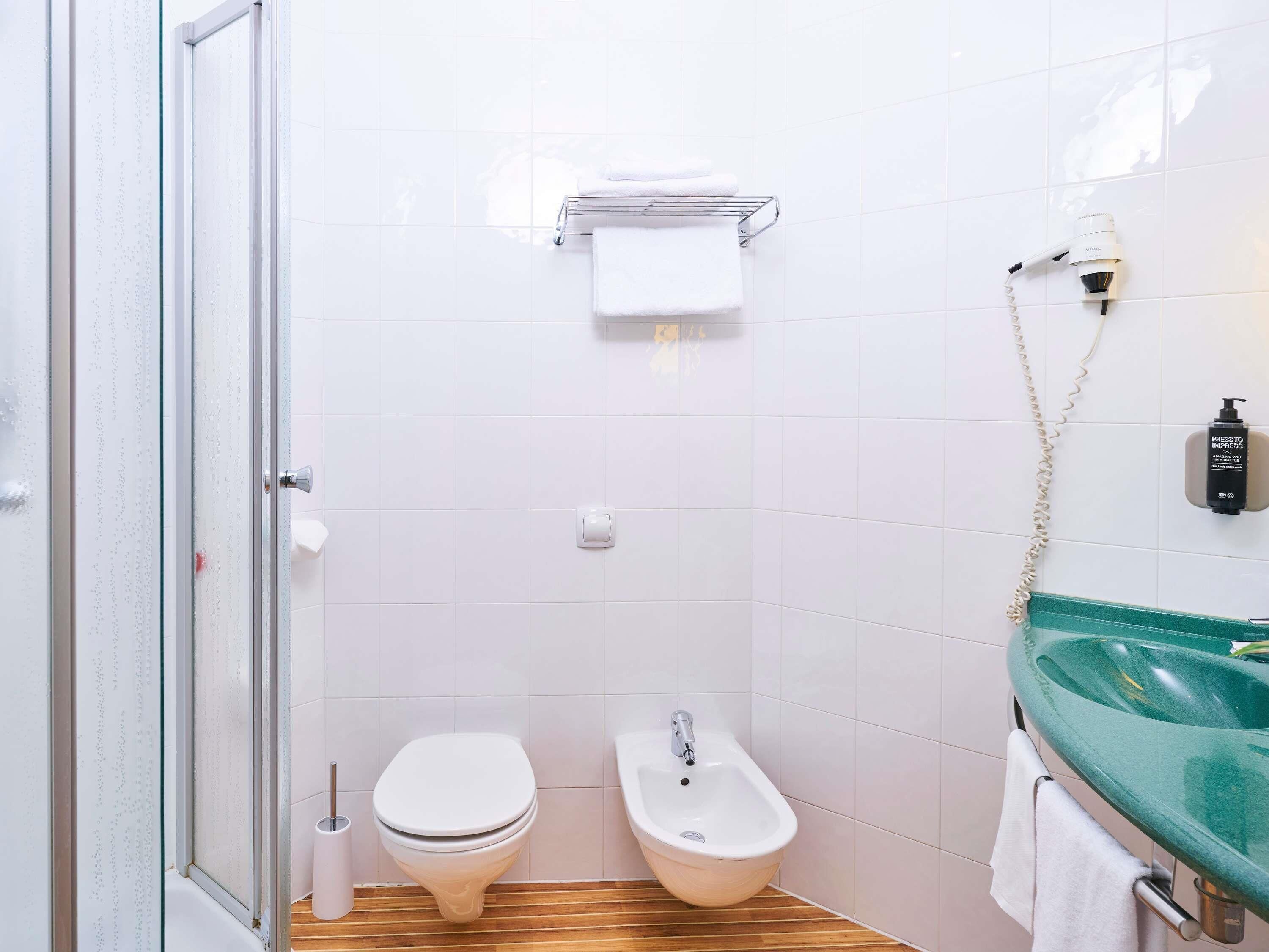 Un bany amb una porta de vidre per a la dutxa, un vàter, un bidet, un lavabo amb una mànega verda i una barra per penjar tovalloles.