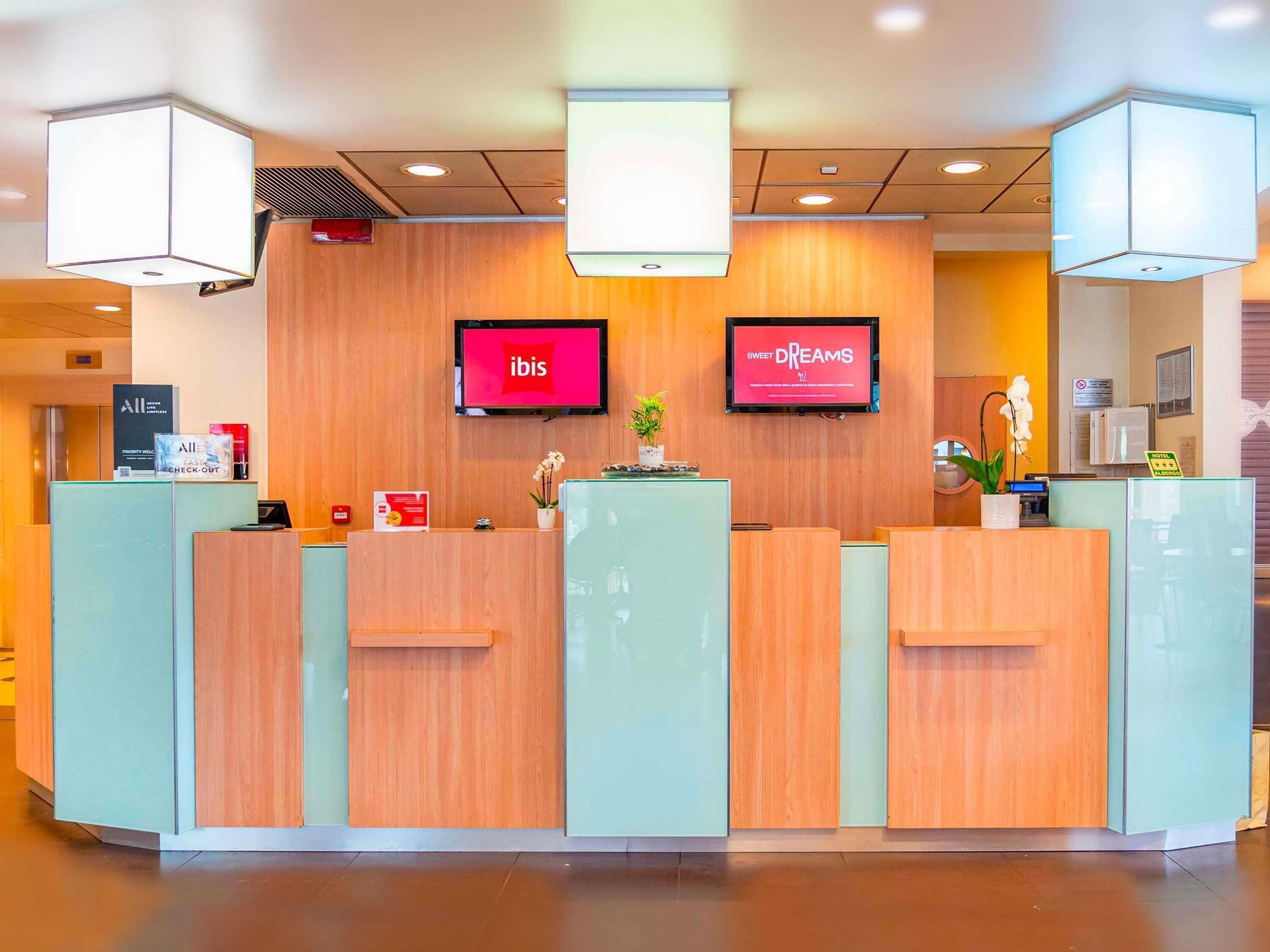 Un mostrador de recepción de hotel con dos pantallas digitales mostrando los logotipos de 'ibis' y 'Sweet Dreams', un letrero de check-out prioritario y un letrero de 'Hotel Check-In'.