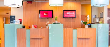 Un mostrador de recepción de hotel con dos pantallas digitales mostrando los logotipos de 'ibis' y 'Sweet Dreams', un letrero de check-out prioritario y un letrero de 'Hotel Check-In'.