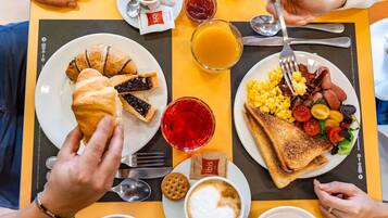 Mesa de café da manhã com croissants, torradas, ovos mexidos, bacon, tomates e uma xícara de café.