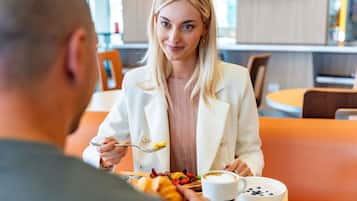A person seated at a table with a meal, in a modern interior with orange seating and a television in the background.
