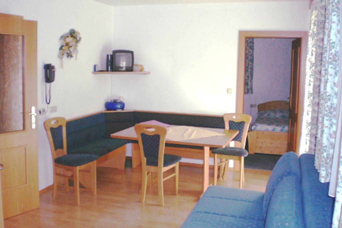 Ein kleiner, gemütlicher Raum mit einem Esstisch, Stühlen, einer Couch, einem Bett und einem Telefon an der Wand.