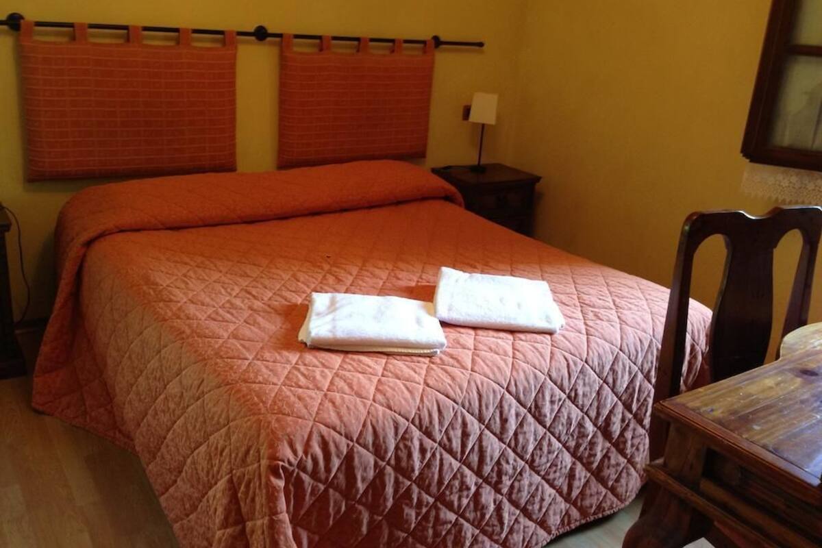 Un lit bien fait, recouvert d’une courtepointe, avec deux oreillers et deux serviettes pliées posées sur le lit. Une petite table de chevet avec une lampe se trouve à droite, ainsi qu’une chaise.