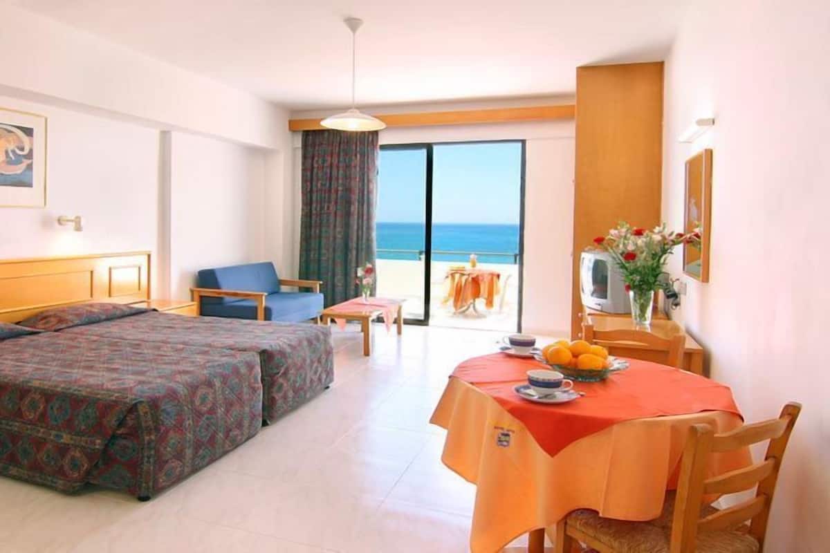 Habitación de hotel con dos camas, una mesa de comedor con mantel naranja, televisor y vista al mar.
