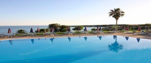 A clear blue pool with lounge chairs and umbrellas, a palm tree, and a view of the sea. - Corallia Beach Hotel Apartments (Pegeia)