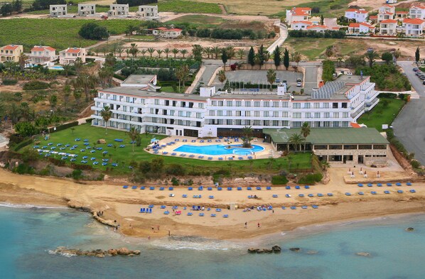 An aerial view of a resort with a large pool, multiple beachside umbrellas, and a beachfront. - Corallia Beach Hotel Apartments (Pegeia)