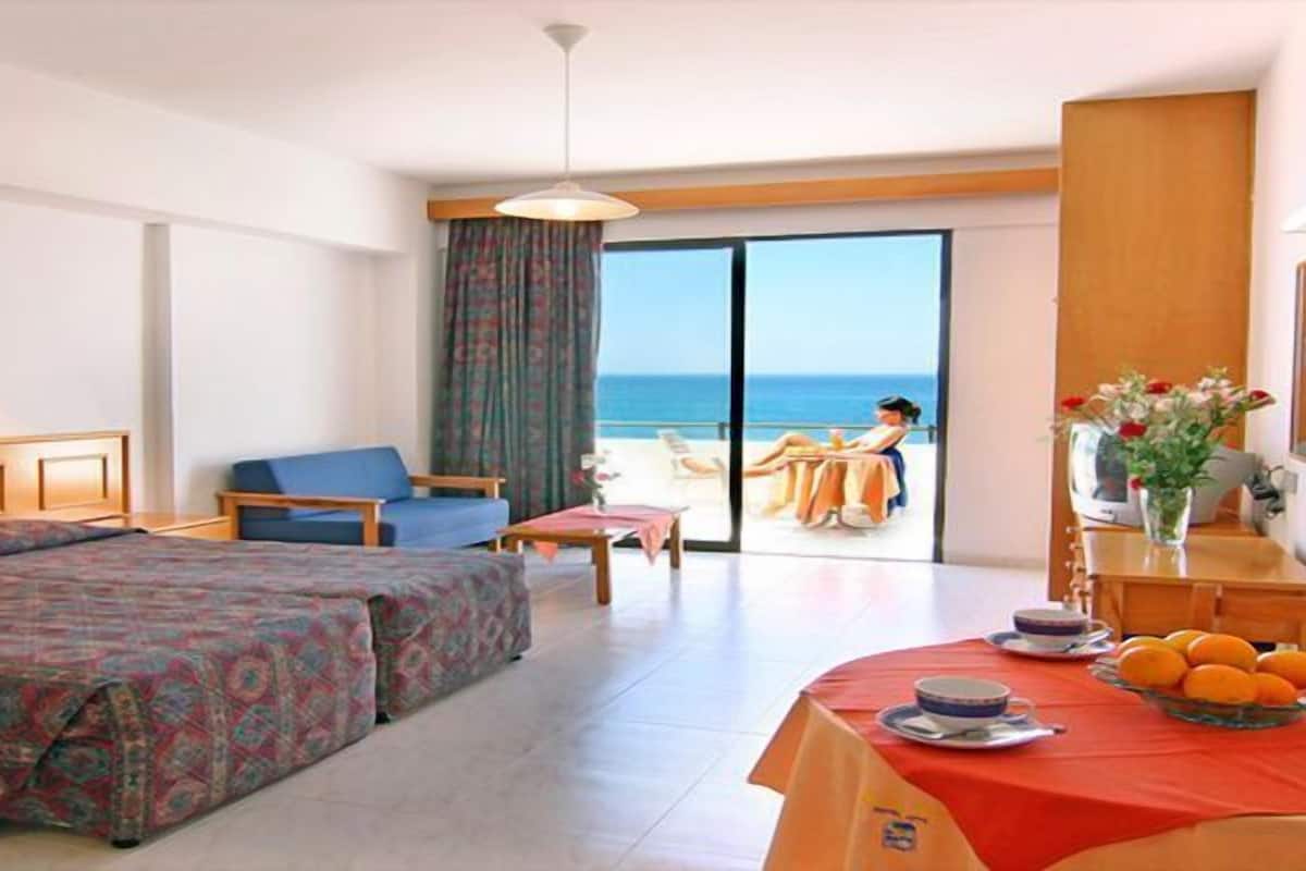 Una habitación de hotel con una cama grande, un sofá, una mesa de comedor con mantel rojo y vistas al mar a través de un ventanal amplio.