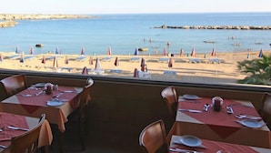 Un restaurante frente al mar con mesas dispuestas para comer, con vista a una playa con sombrillas y gente.