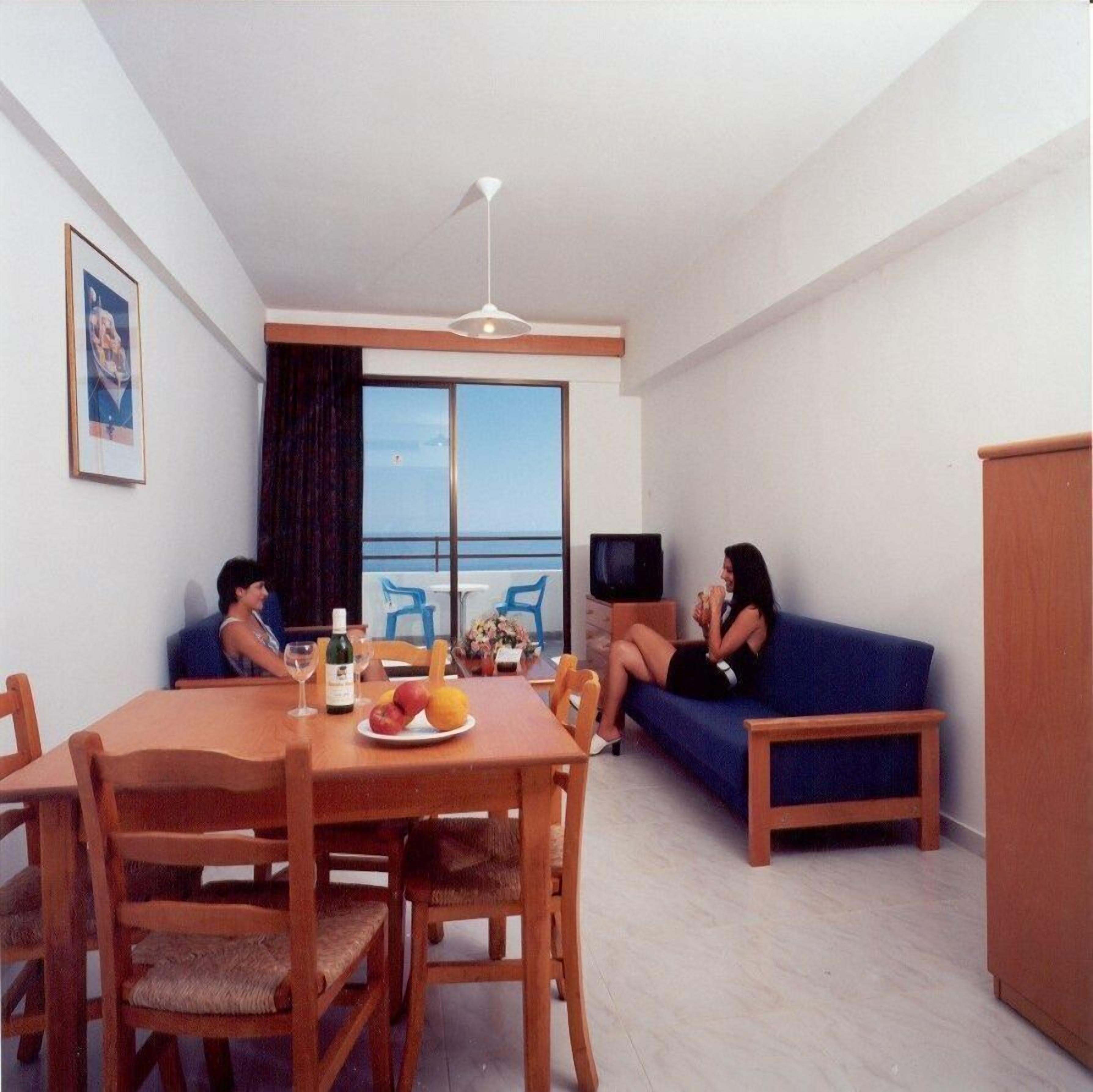 A modern living room with a dining area, a sofa, a TV, and a balcony with a view of the sea.