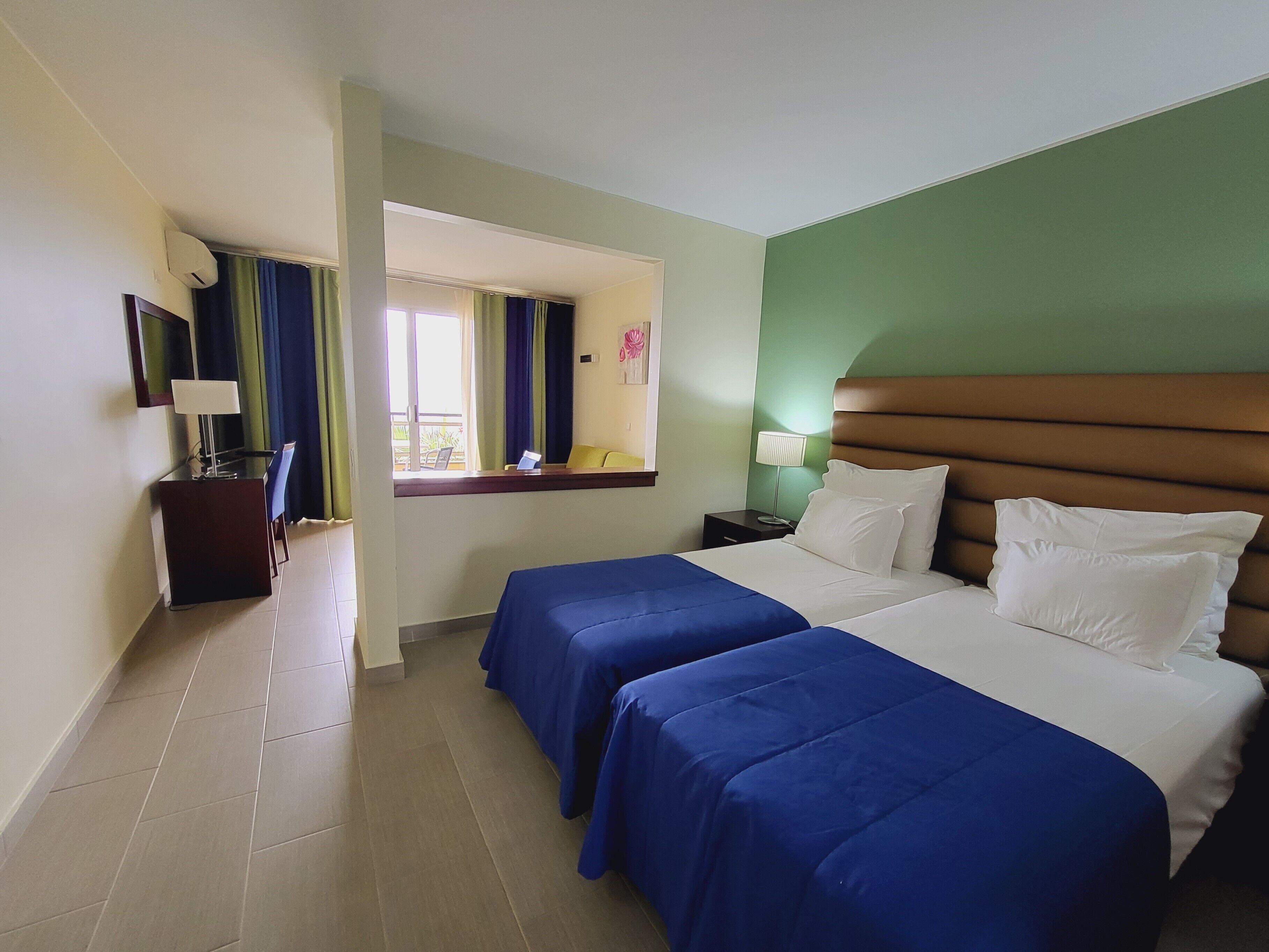 A hotel room with two beds, a desk with a chair, a desk lamp, a nightstand with a lamp, and a window with curtains.
