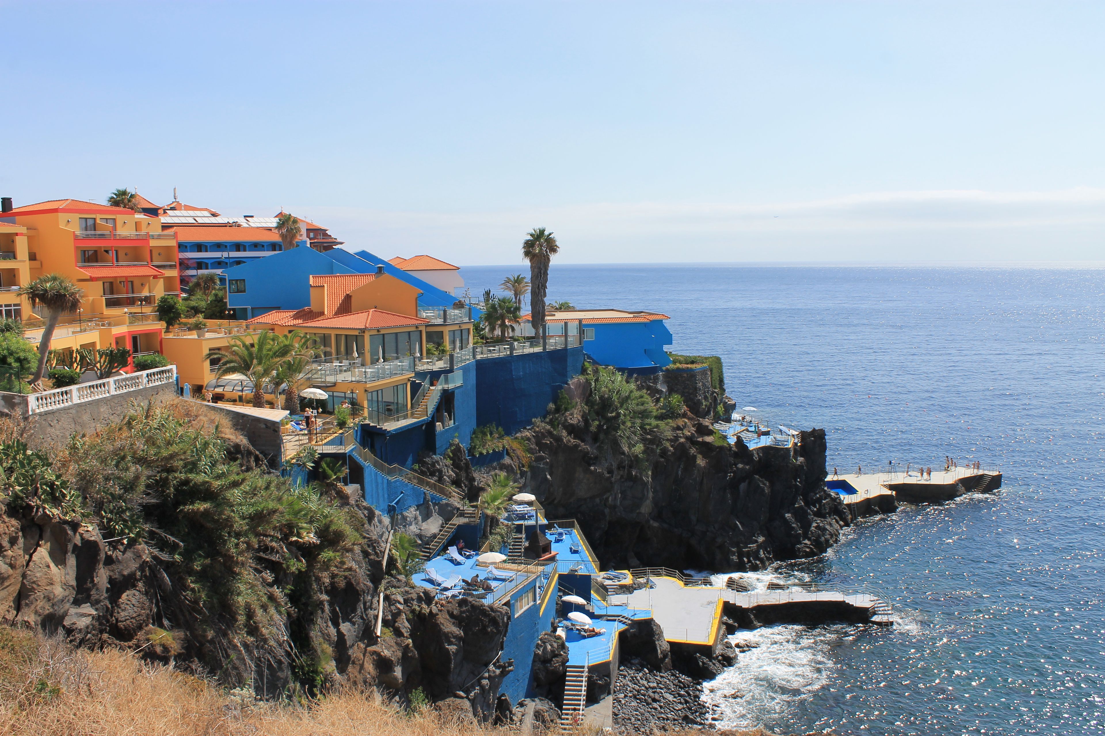 A coastal hotel with multiple balconies overlooking the sea, a pool area, and a rocky shoreline.