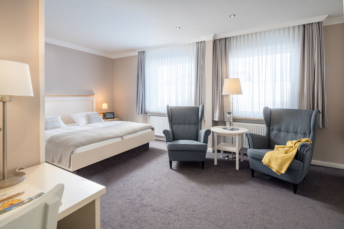 Una habitación de hotel con una cama, dos sillones, una mesita y una lámpara.