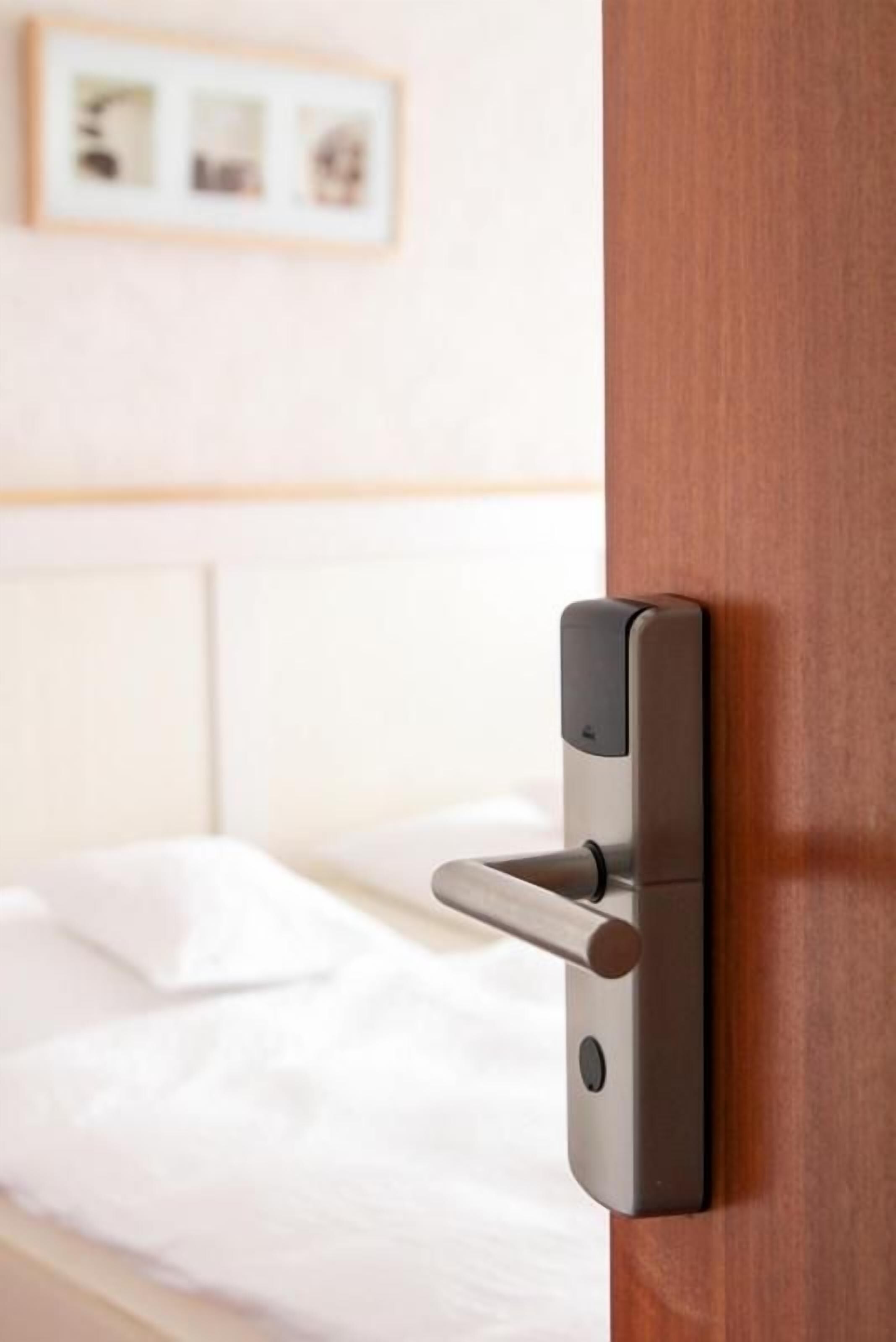 Une poignée de porte moderne, dotée d’un levier et d’un trou de serrure, faisant partie d’une pièce où se trouve un lit blanc et un tableau encadré au mur.