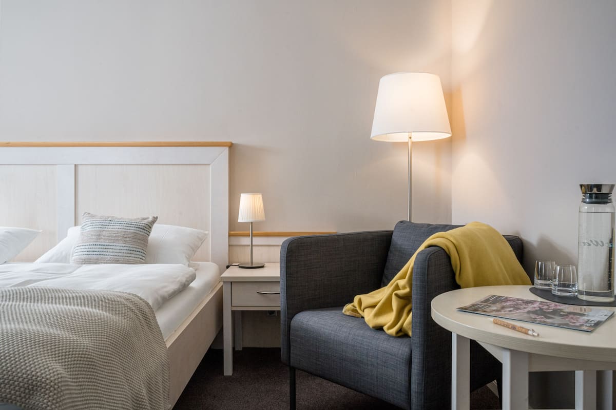 Habitación de hotel con cama, mesita de noche, lámpara, silla y una pequeña mesa con vasos y una revista.