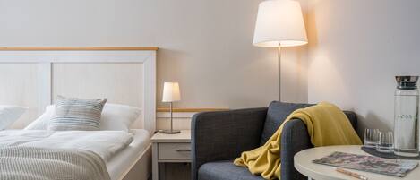 Habitación de hotel con cama, mesita de noche, lámpara, silla y una pequeña mesa con vasos y una revista.