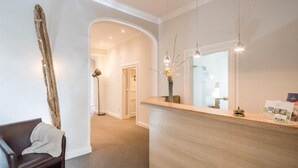 A reception area with a wooden reception desk, a dark leather bench, a tall vase with flowers, and a mirror reflecting the interior. - AKZENT Hotel Dorn (Büsum)